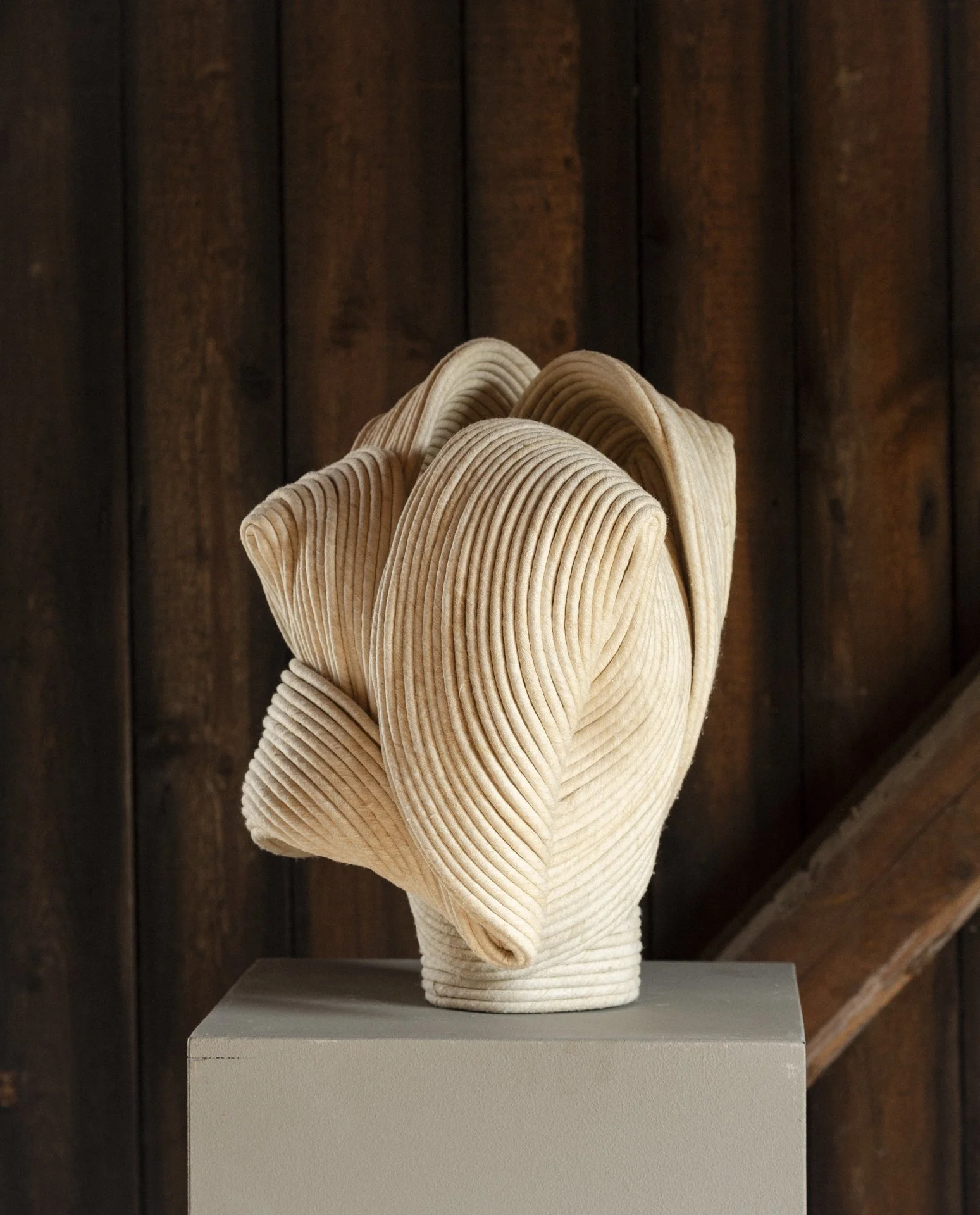 Sculpture resembling a flower, made of folded, ribbed fabric, displayed on a pedestal against a wooden background.