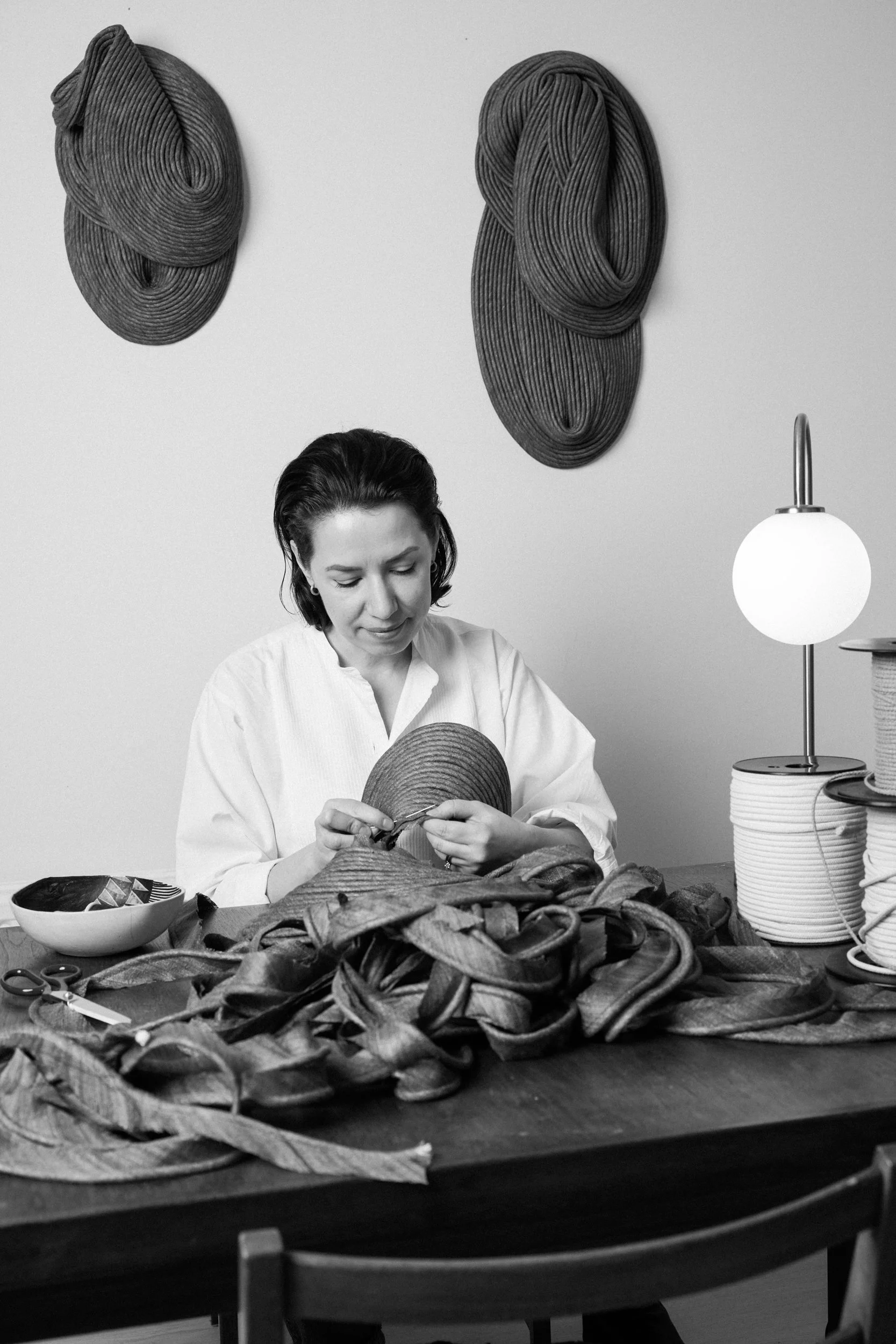 A woman sitting at a table sewing fabric, with large woven textile art pieces hanging on the wall behind her and spools of thread on the table.