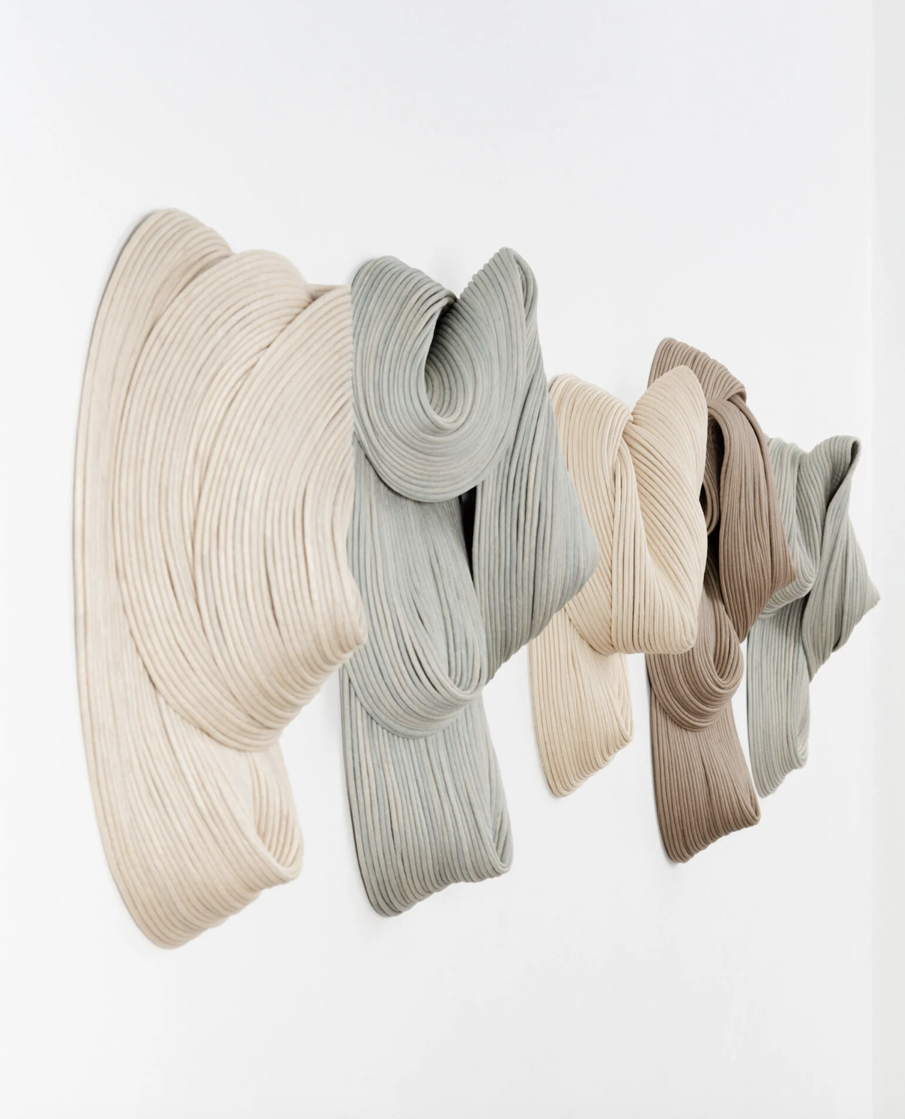 An abstract art installation of five three-dimensional, undulating, ribbon-like sculptures in neutral colors, mounted on a white wall.