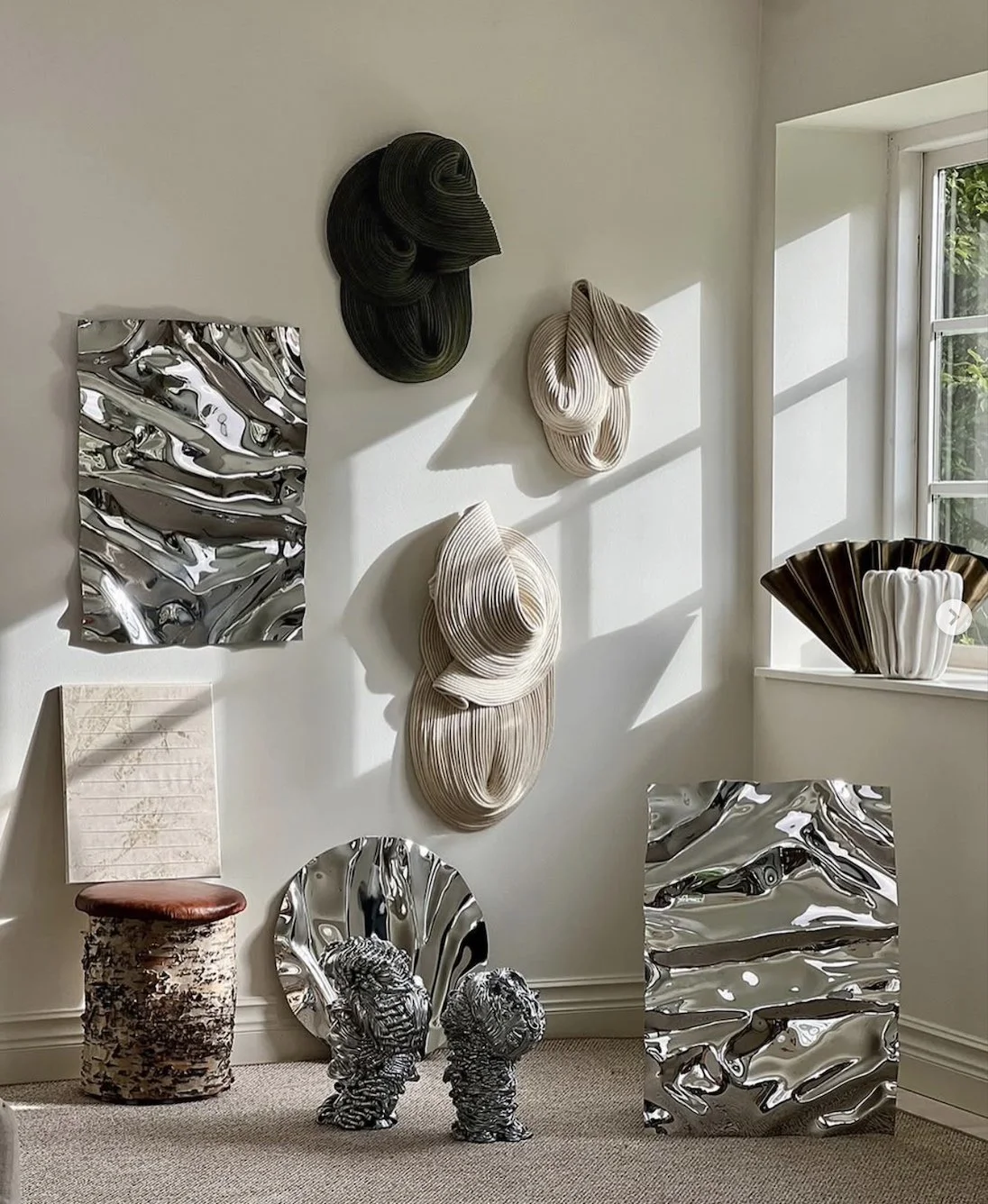 Interior display with wall-mounted abstract sculptures, metallic and ceramic art pieces, including fabric-like, shiny, and textured materials, placed in natural daylight near a window.