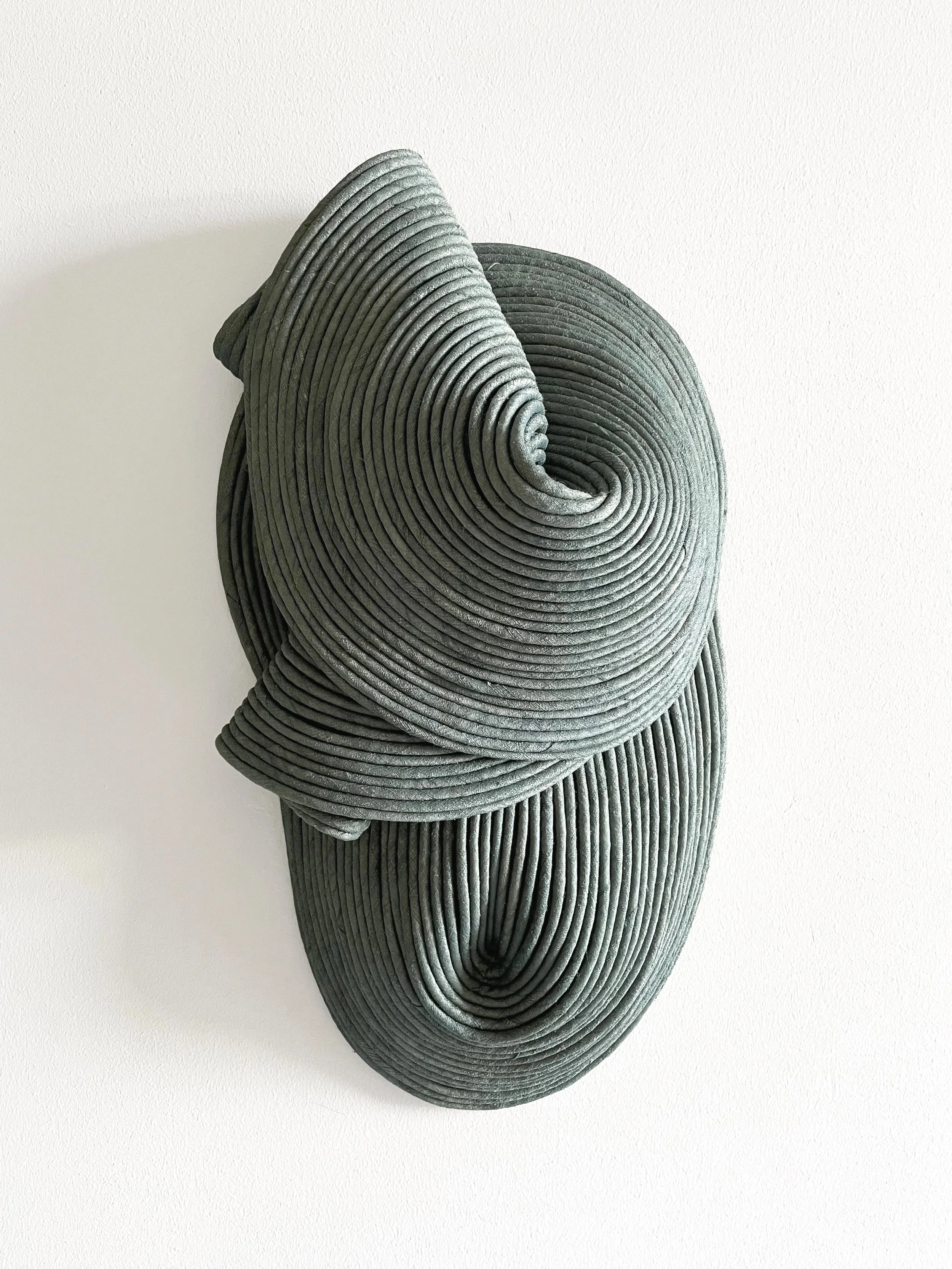 An abstract sculpture made of concentric, folded, spiral-shaped gray fabric or material mounted on a white wall.