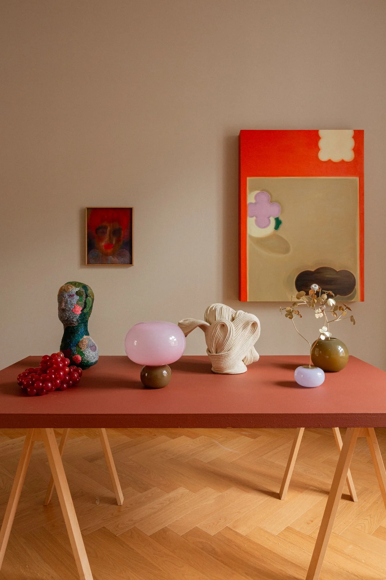 A minimalistic art display showcases a pink tabletop with six variously shaped vases and sculptures, against a beige wall adorned with three abstract paintings.