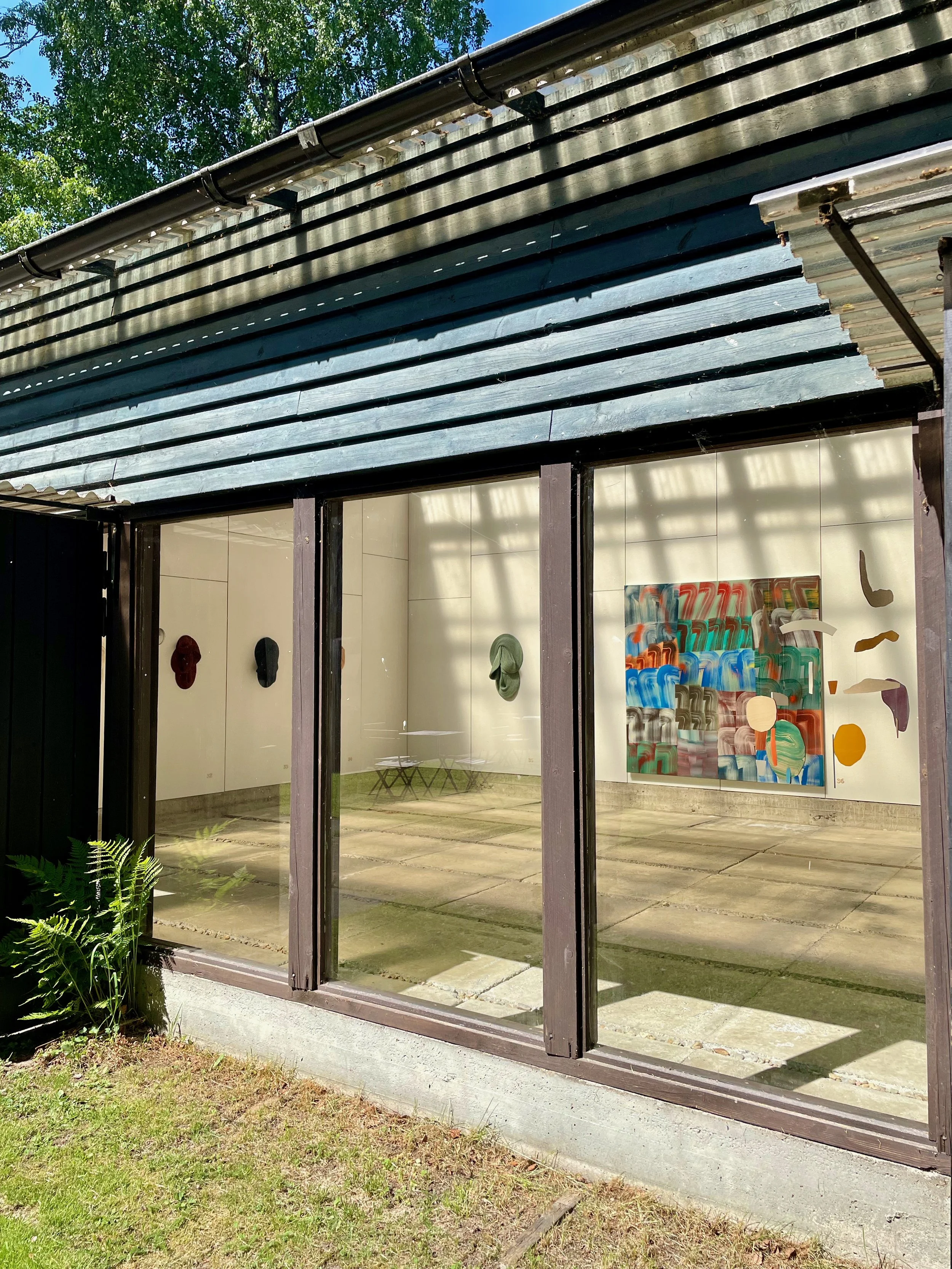 View of an art gallery with large glass windows, artist's colorful abstract paintings on the white walls inside, and outdoor greenery including a fern plant near the window.