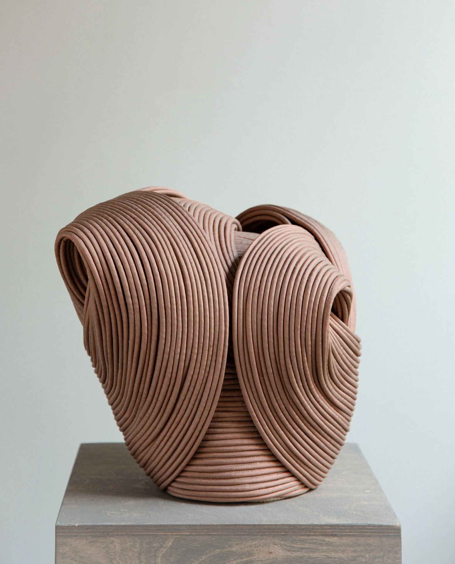 Contemporary ceramic sculpture resembling an abstract human head, composed of pinkish-brown ridged clay coils, displayed on a grey pedestal against a plain wall.