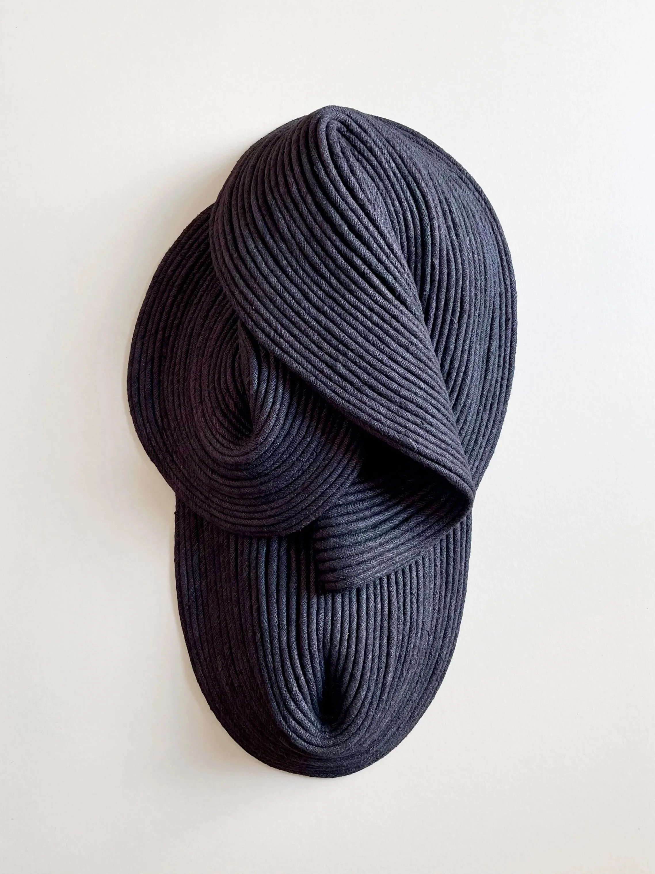 A wall sculpture made of coiled black cord or fabric, arranged in an abstract, flowing shape on a white wall.