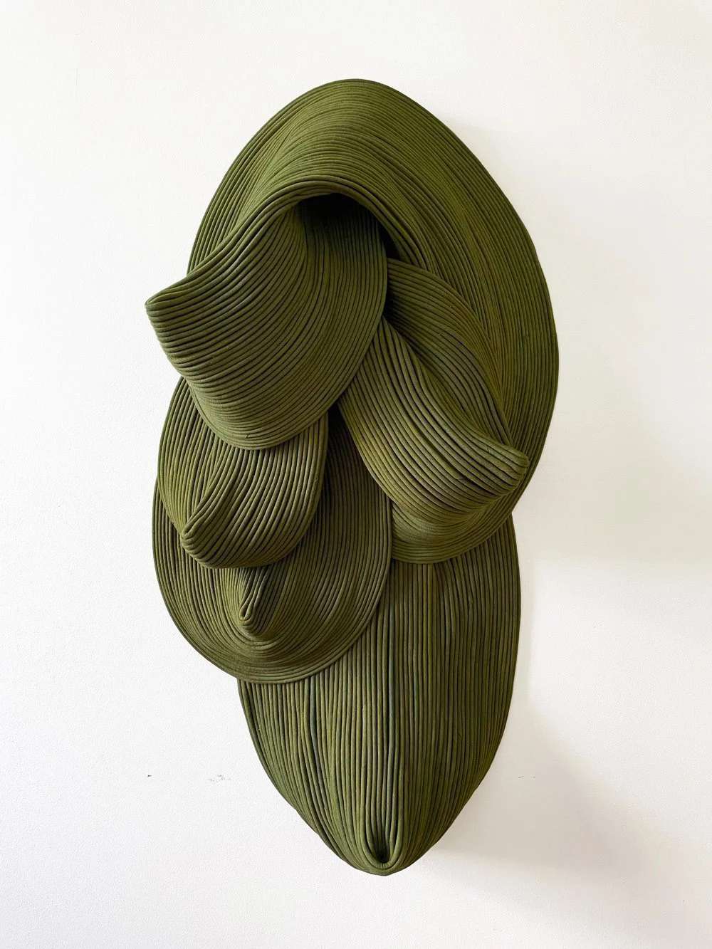 An abstract sculpture made of stacked, concentric green forms resembling a stylized face or mask, mounted on a white wall.