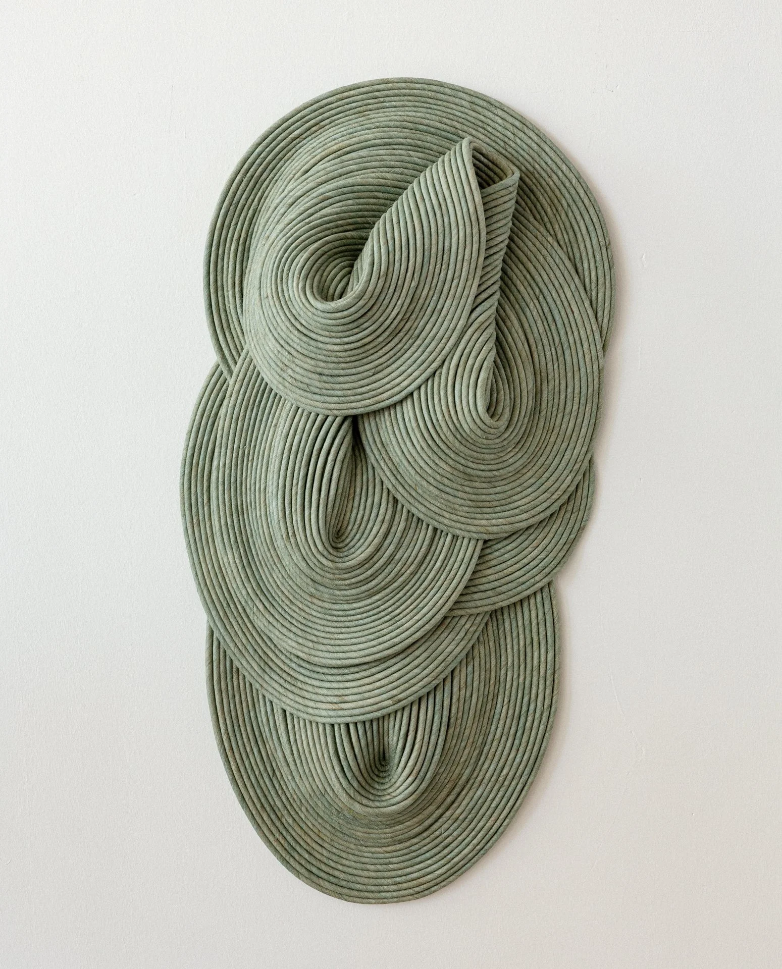 A modern art wall sculpture made of overlapping, circular, gray, coil-like elements arranged vertically against a plain white wall.