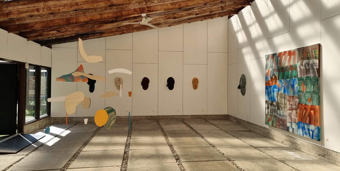 Interior of an art gallery with hanging abstract sculptures and a colorful abstract painting on the wall, sunlight streaming through windows.