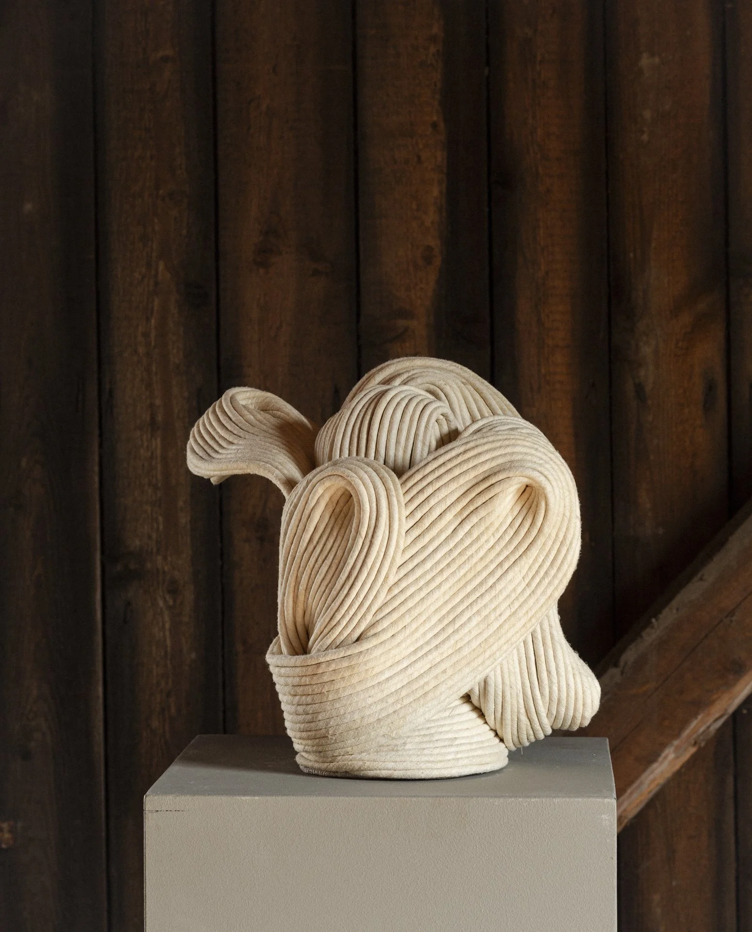 Artistic sculpture made of off-white, ribbed, cord-like material, resembling an abstract face or head, displayed on a gray pedestal against a wooden wall background.