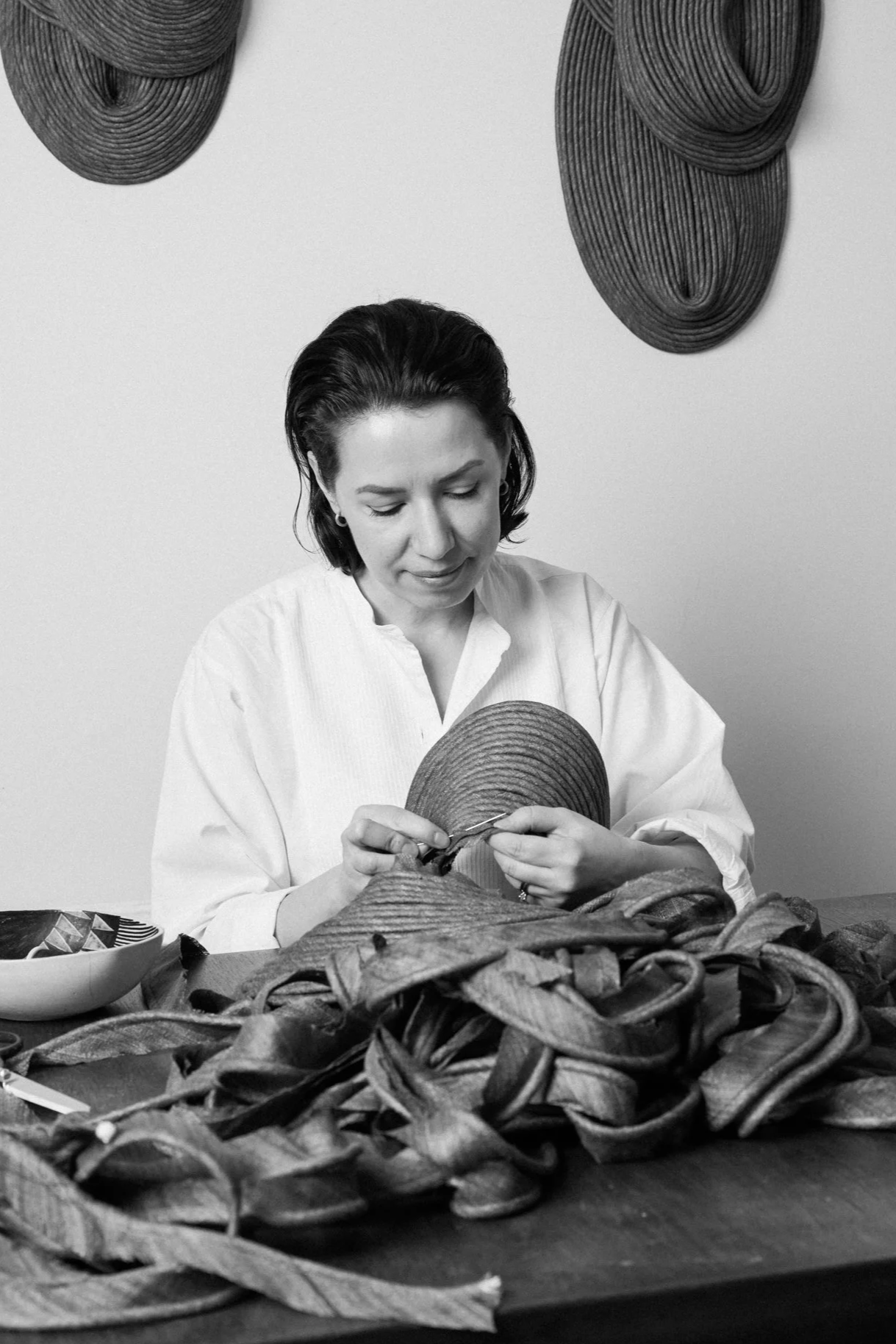 A woman with short dark hair, wearing a white shirt, is working with woven straw or rattan material, possibly making a craft or basket. There are decorative woven straw pieces hung on the wall behind her.