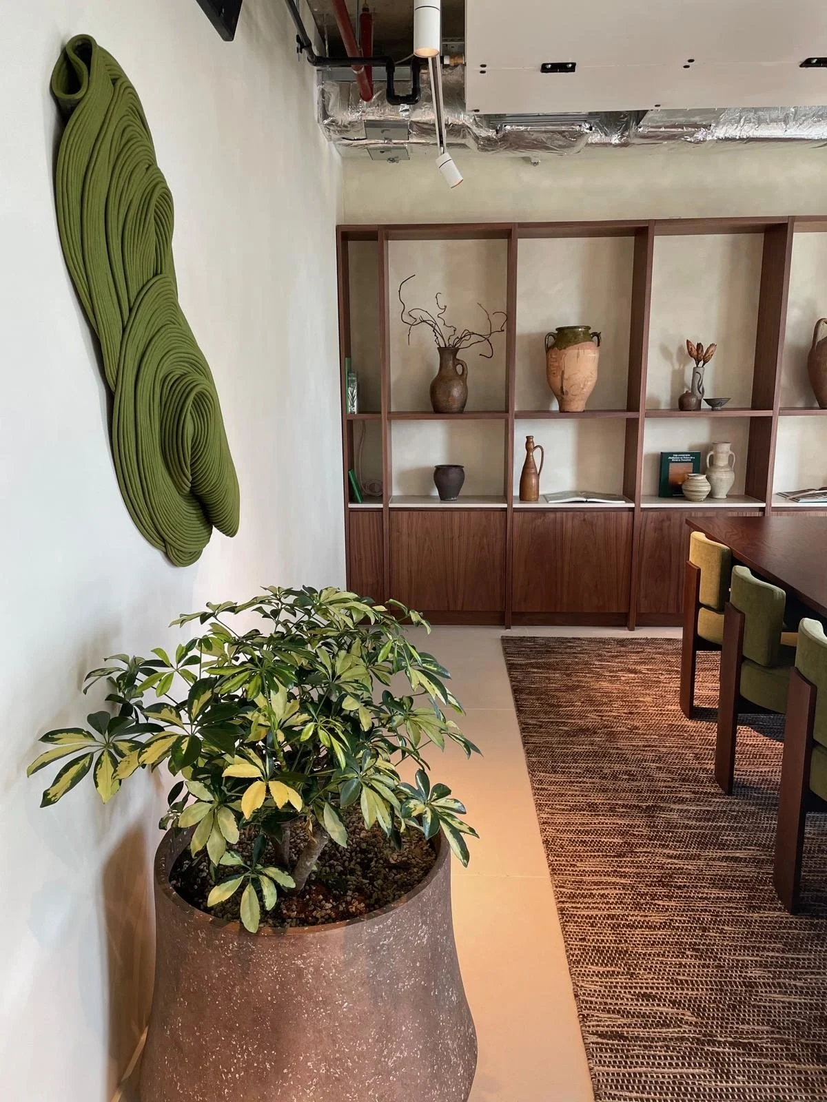 A room with a large potted plant in the foreground, a wooden bookshelf with decorative vases and books on the back wall, and part of a wooden dining table with green chairs. Green wall art is mounted on the left wall. The ceiling has visible ductwork and a hanging light fixture.