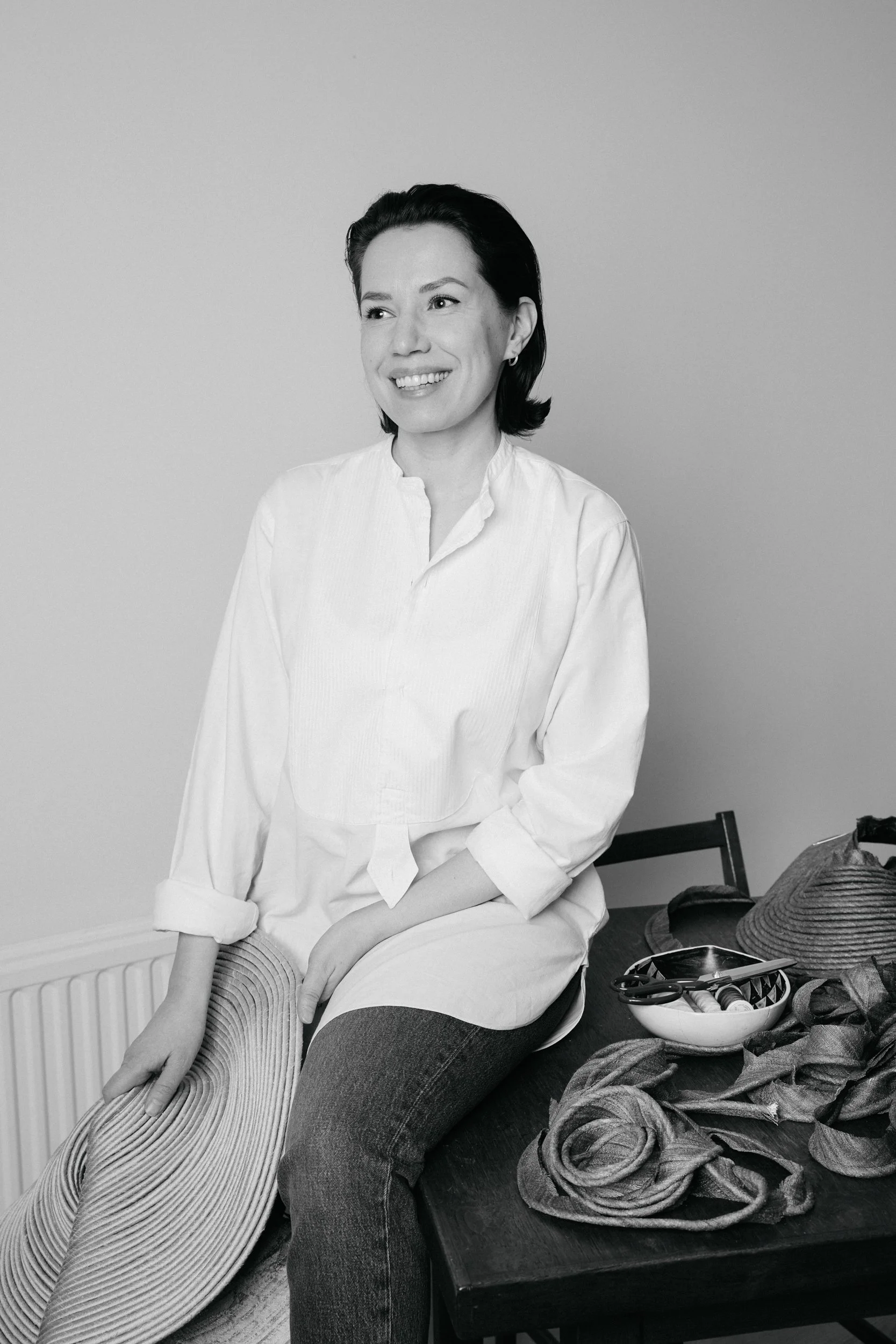 A woman sitting on a table, smiling, with a wide-brimmed hat, scissors, fabric, and other items on the table beside her.
