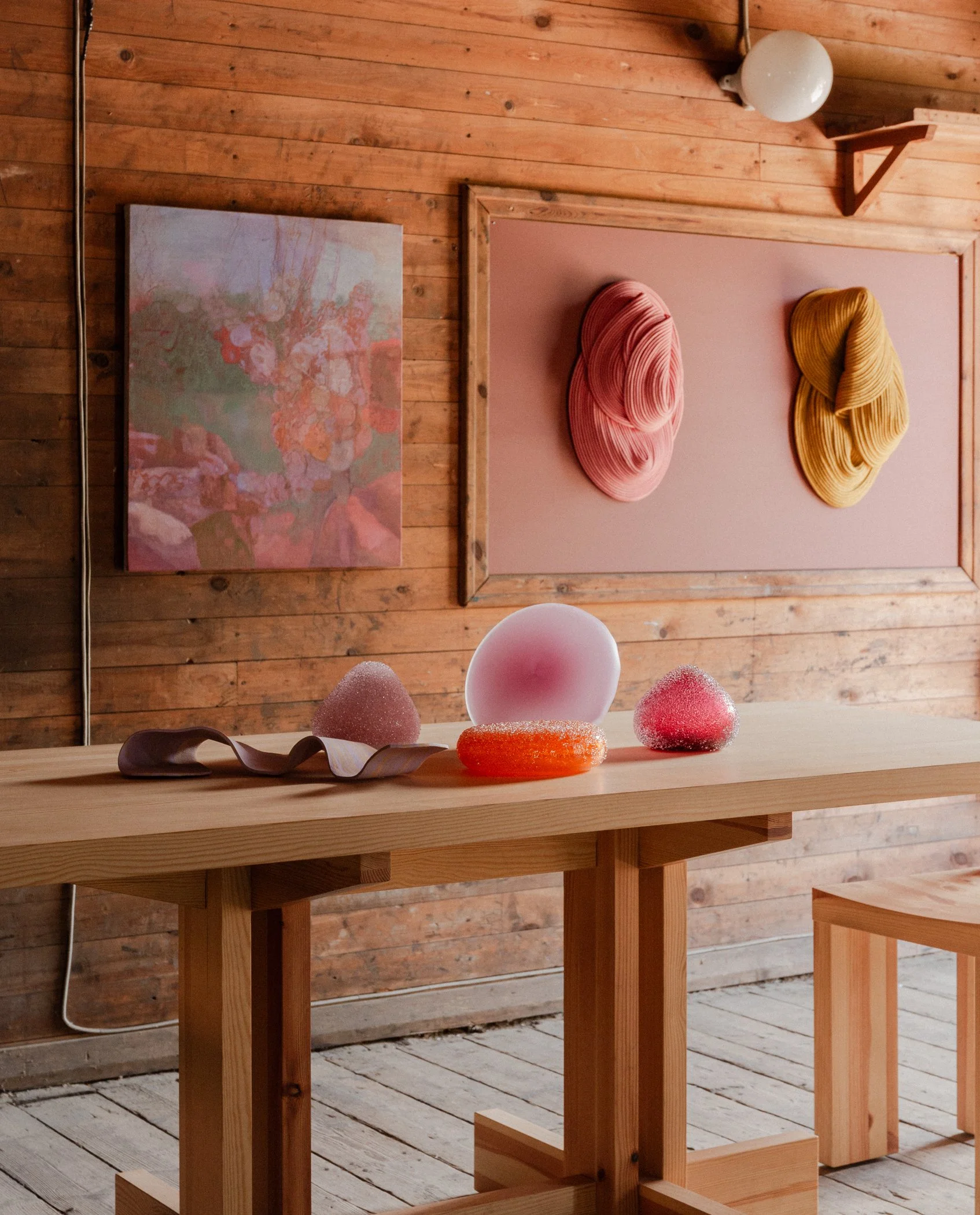 Art gallery with abstract pink and yellow wall sculptures and paintings, wooden table with colorful glass or ceramic objects in a room with wooden walls and floor.