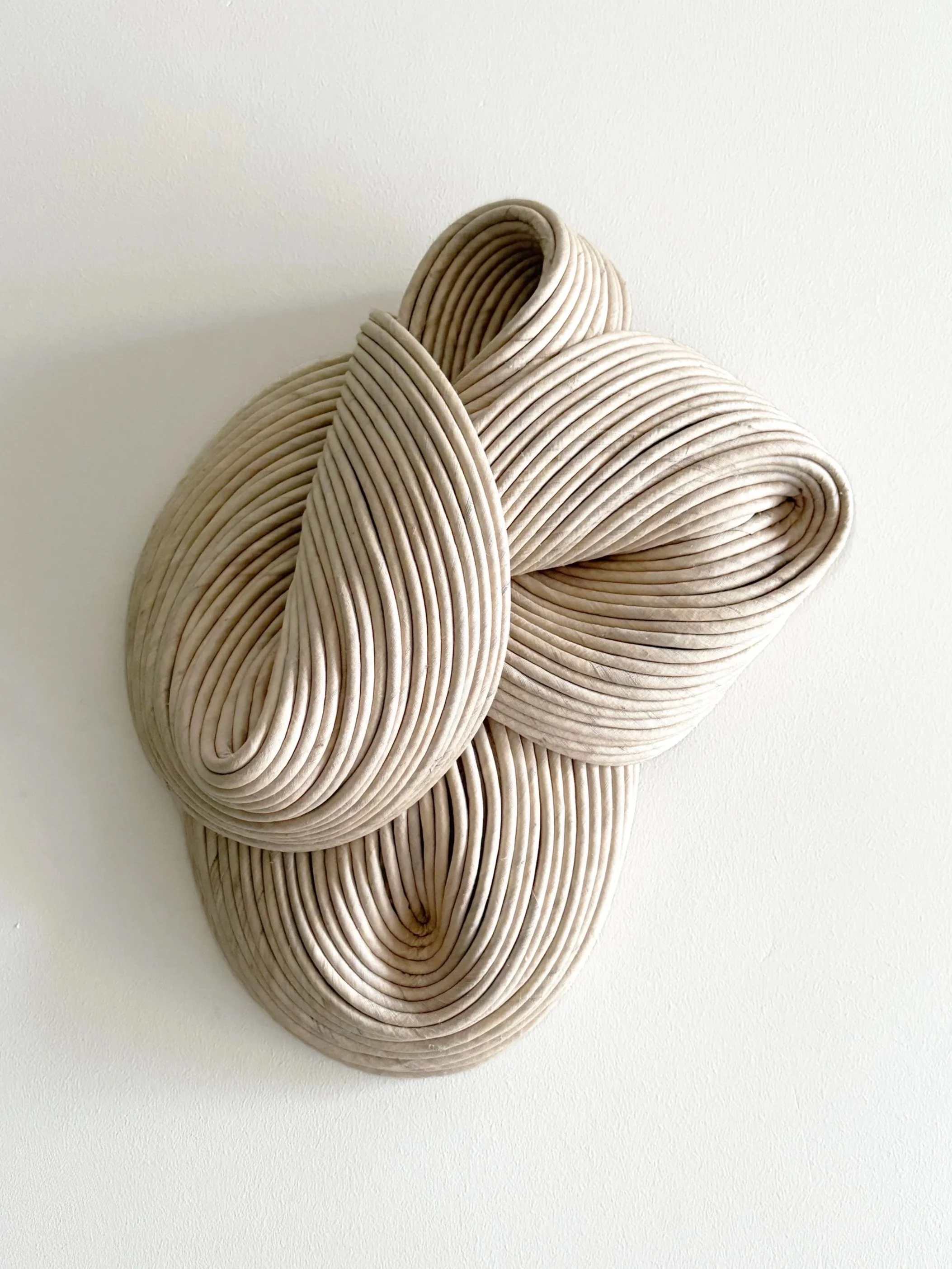 Abstract beige sculpture made of layered, curved, ribbon-like forms mounted on a white wall.