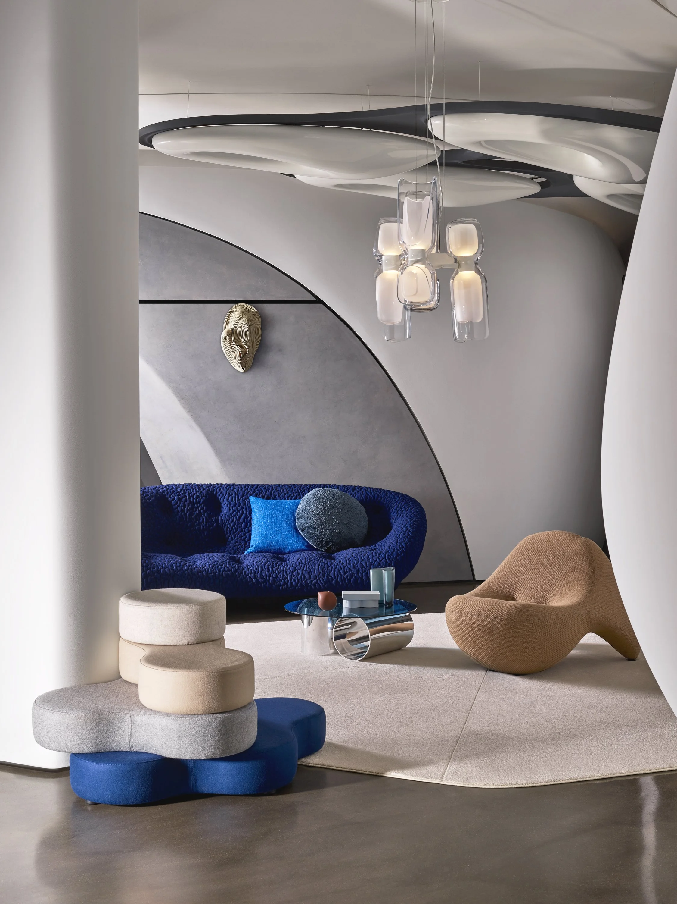 Modern living room with abstract furniture and decor, including a blue textured sofa with blue and round pillows, a beige and brown lounge chair, a layered terrazzo coffee table, and a unique stack of upholstered blocks. The space has a sculptural ceiling installation, contemporary chandelier, curved walls, and a textured gray and white wall background.