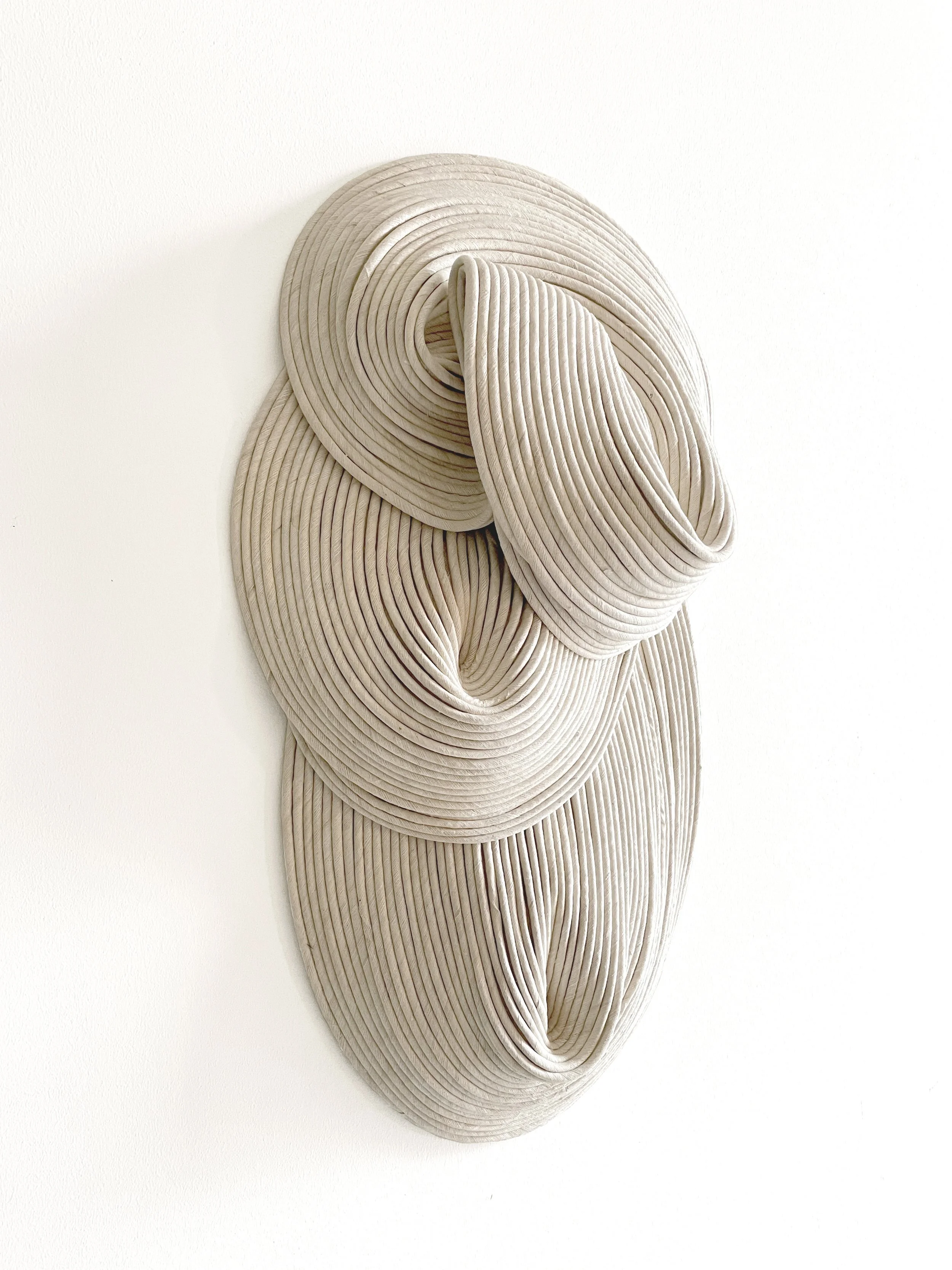 Abstract sculpture made of layered, folded, and curved beige material, resembling flowing fabric or organic form, mounted on a white wall.