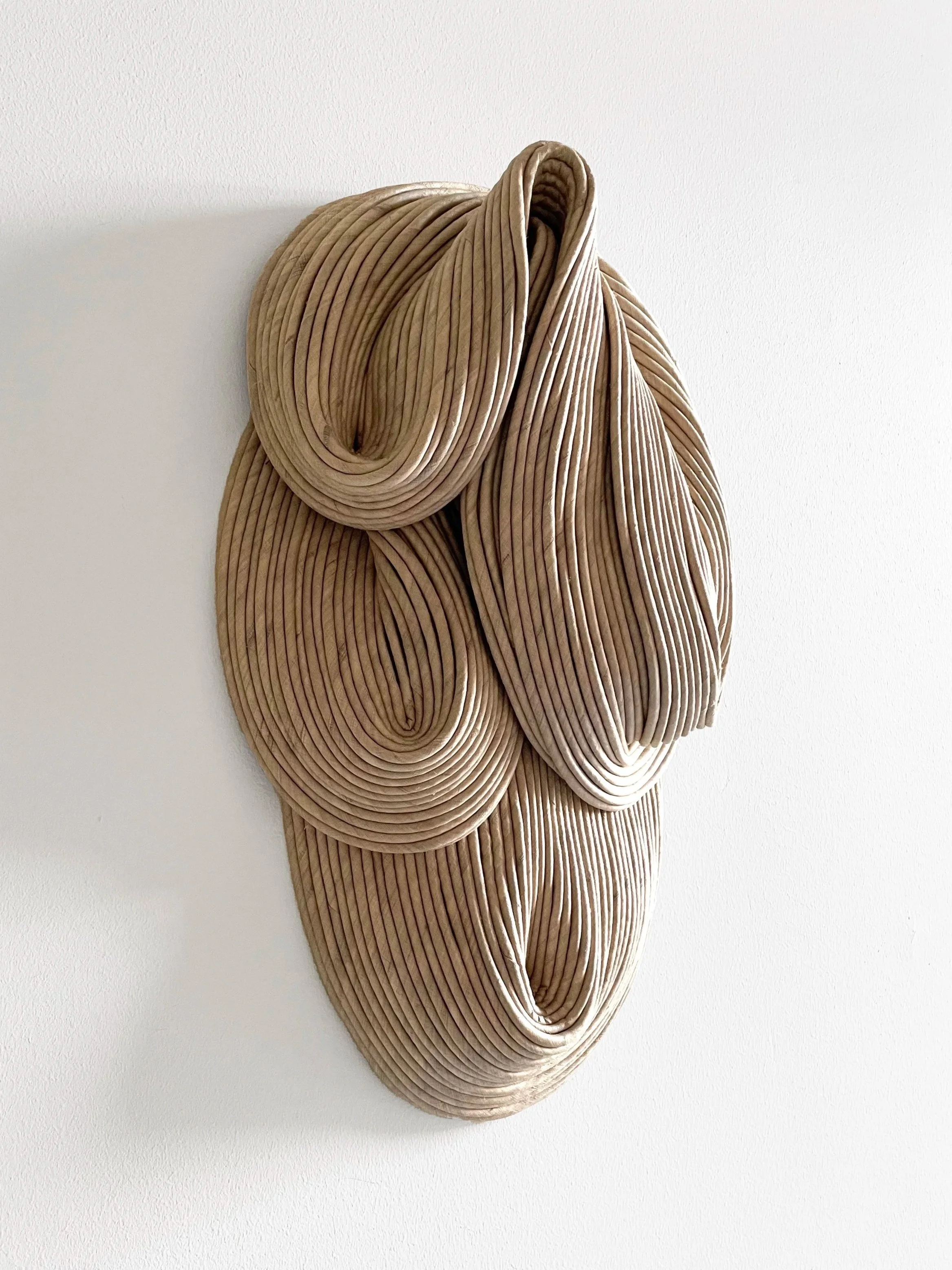 Abstract wall sculpture made of intertwined beige and light brown ridged material resembling fabric or paper, mounted on a white wall.