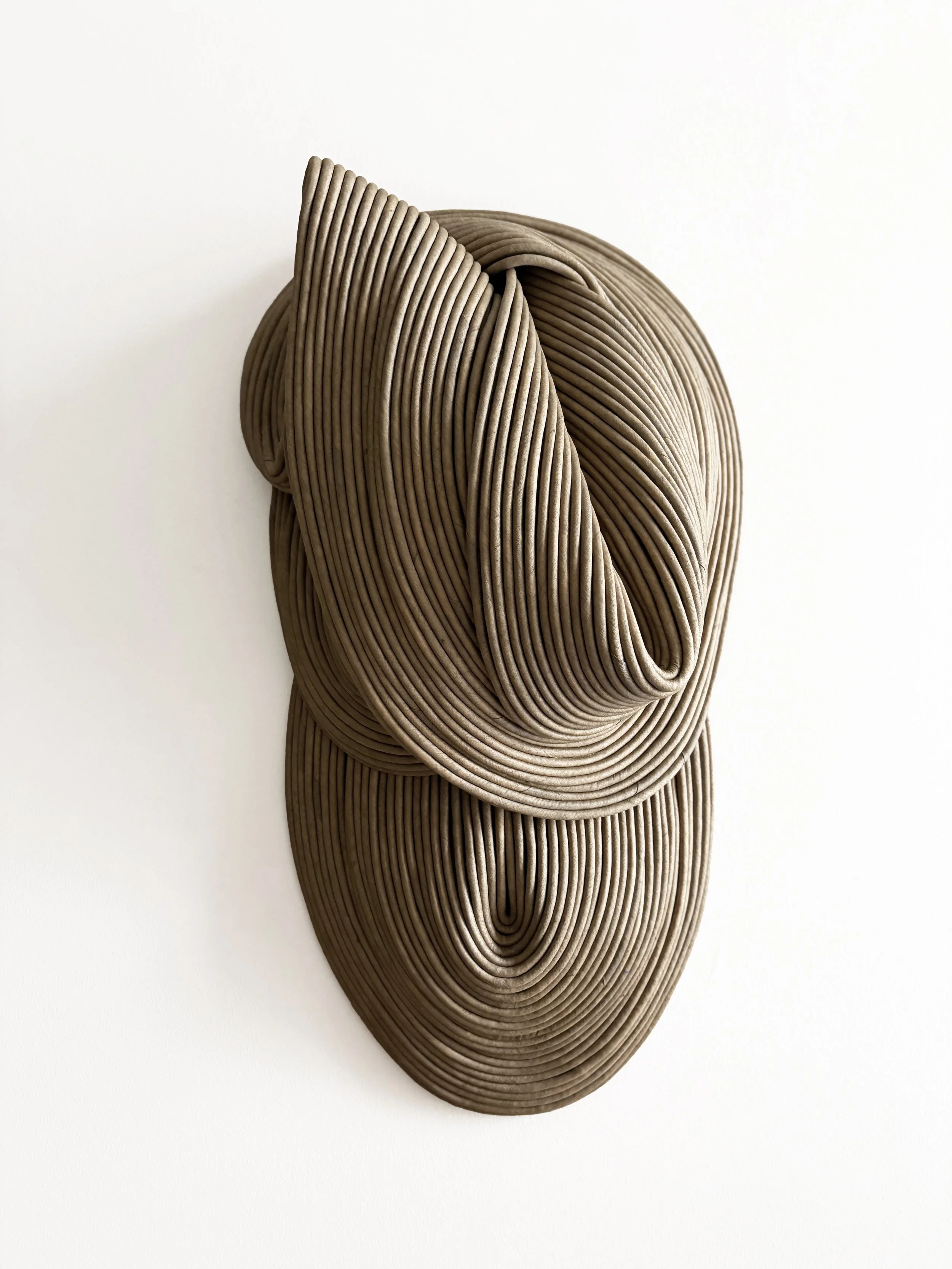 Abstract sculpture composed of layered, curved, ribbed material in shades of beige and brown, resembling a stylized shell or natural formation, set against a plain white background.