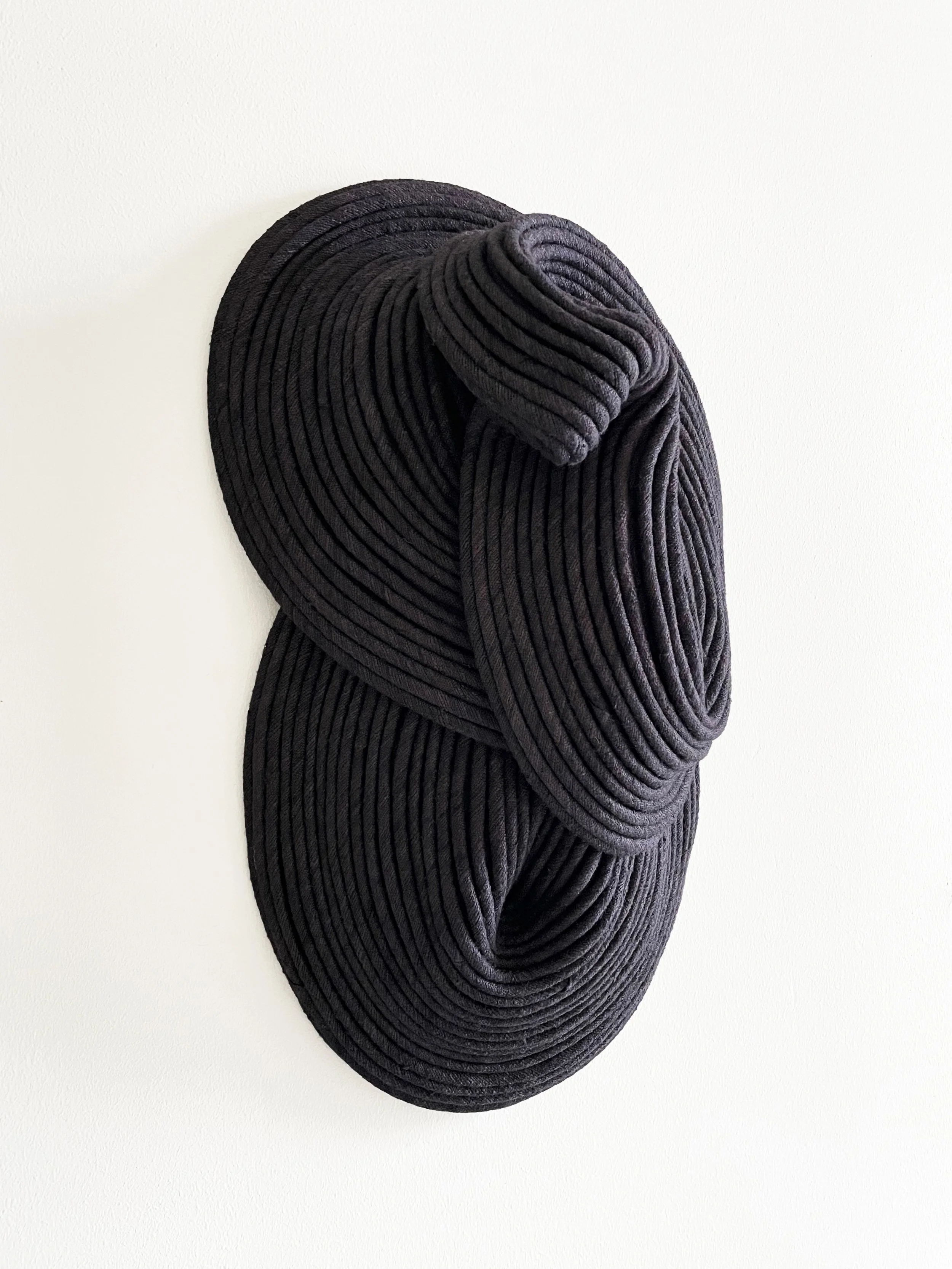 Black and gray textured fabric sculpture with circular ridges, mounted on a white wall.