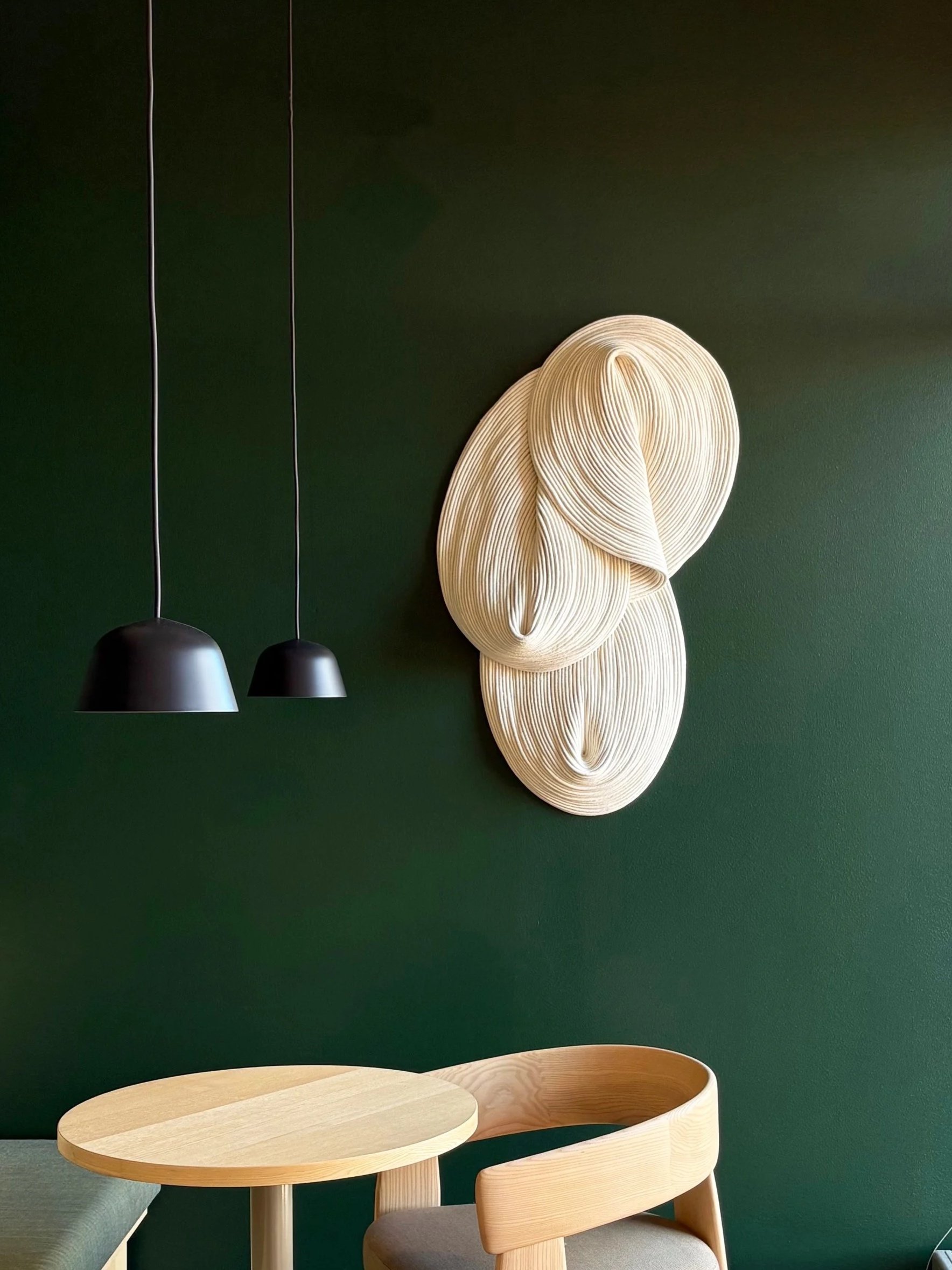 A dark green wall with modern, abstract wooden wall art, two black pendant lights hanging, a round wooden table, and a wooden chair with a beige cushion.