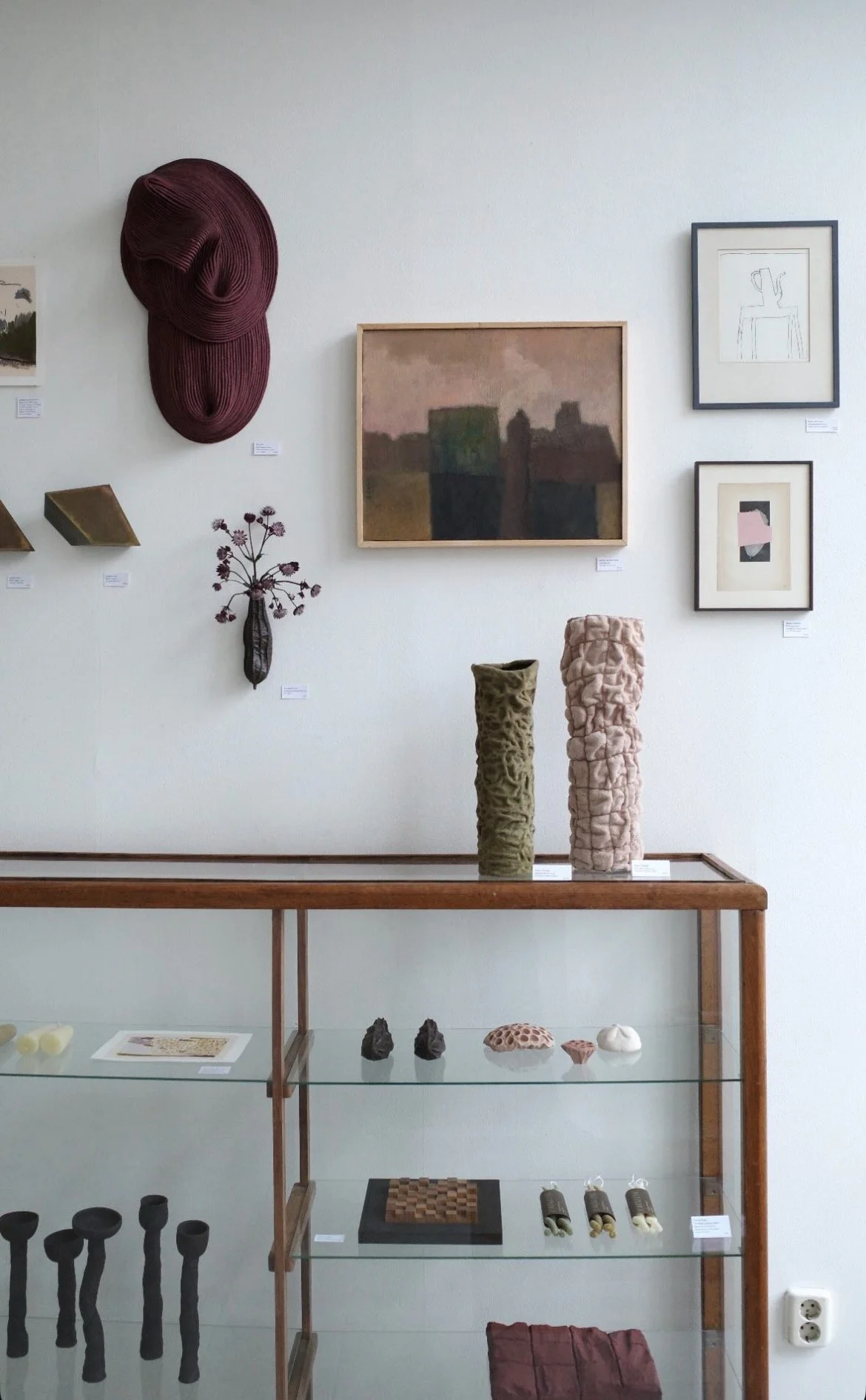 Art display with textured ceramics and sculptures on glass shelves and framed paintings on white wall.