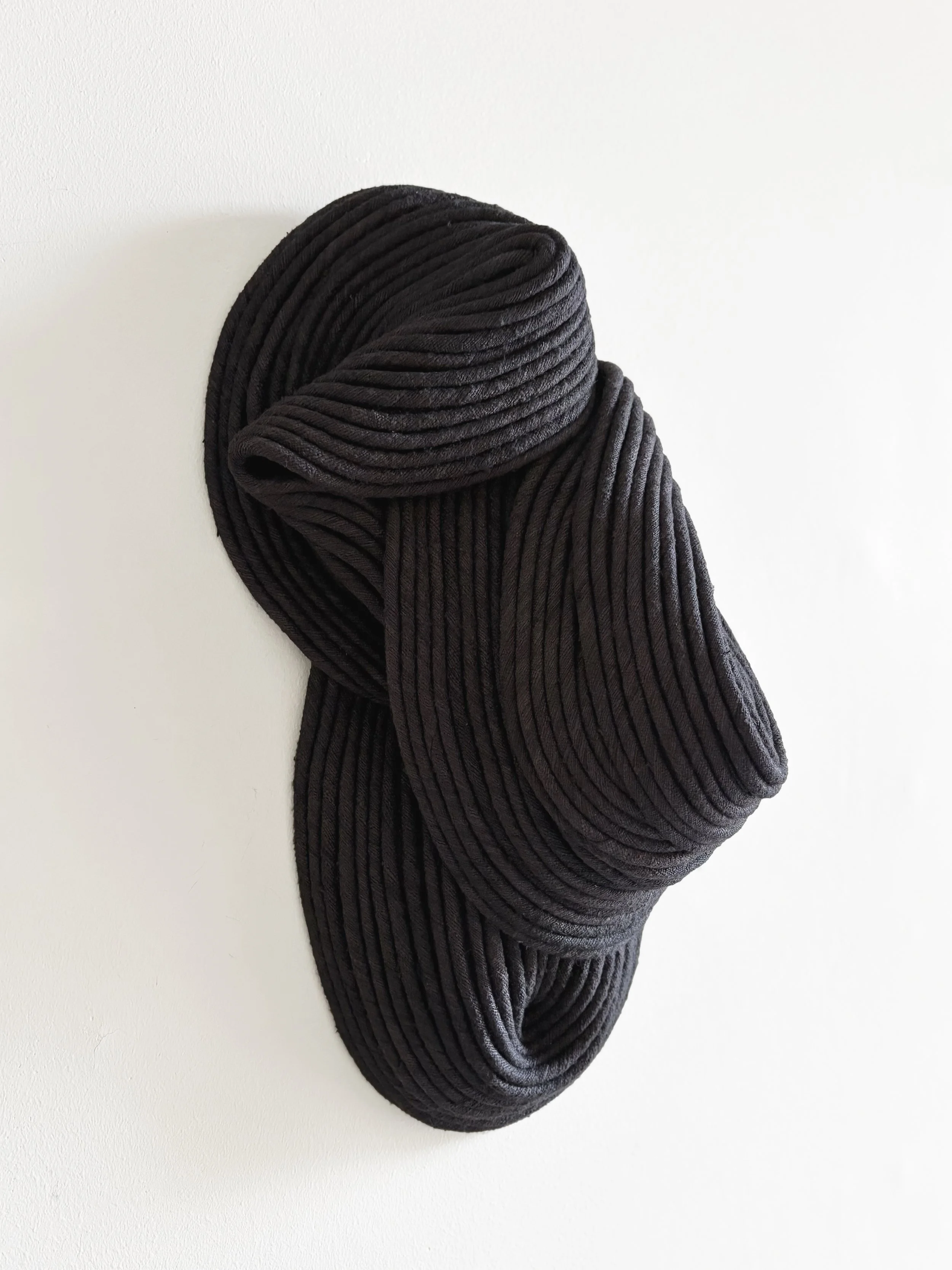 Black textile sculpture with curved, ribbed surfaces mounted on a white wall.