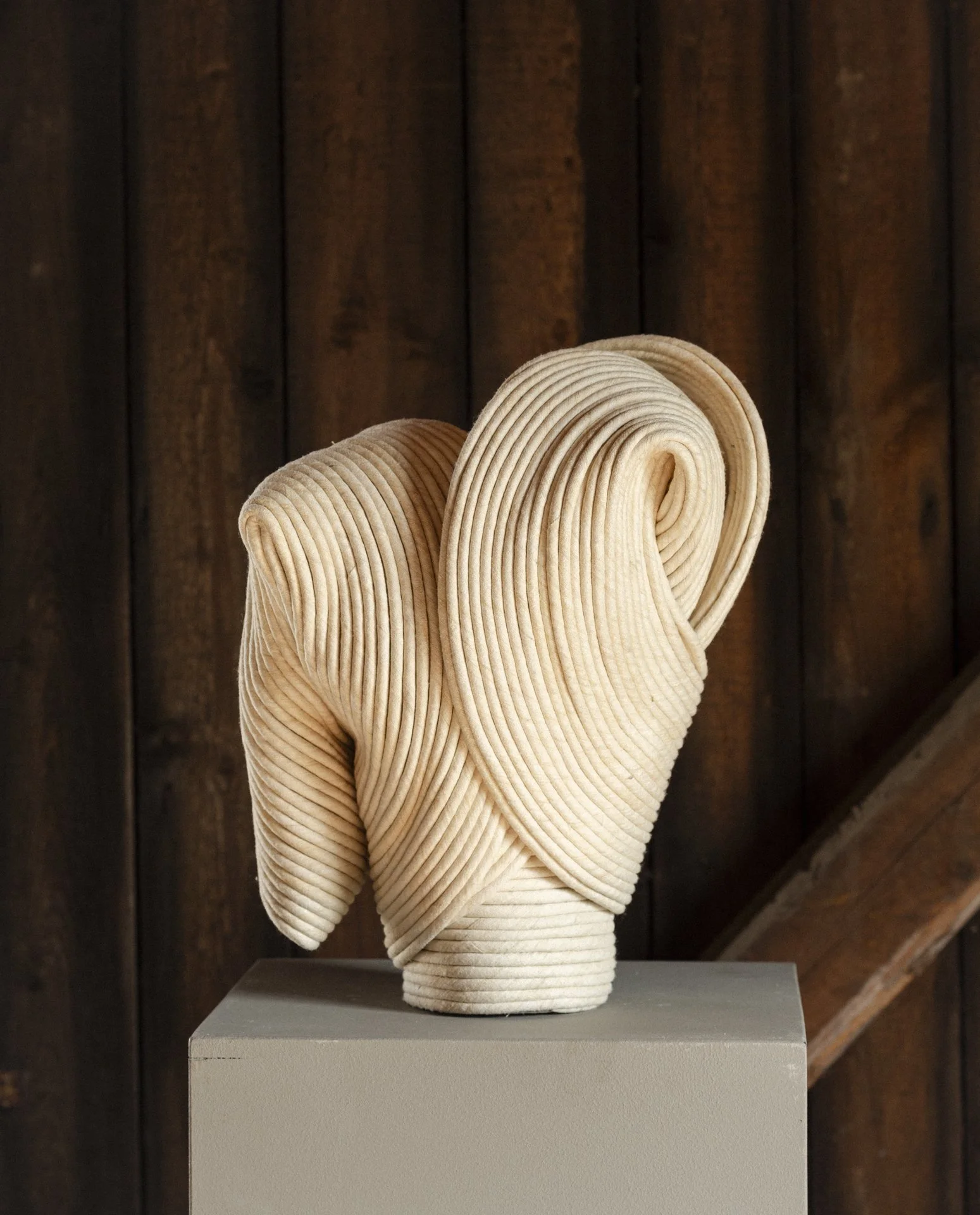 A modern art sculpture of a human face made from stacked, curved, beige-colored cord-like materials, displayed on a gray pedestal against a dark wooden wall.