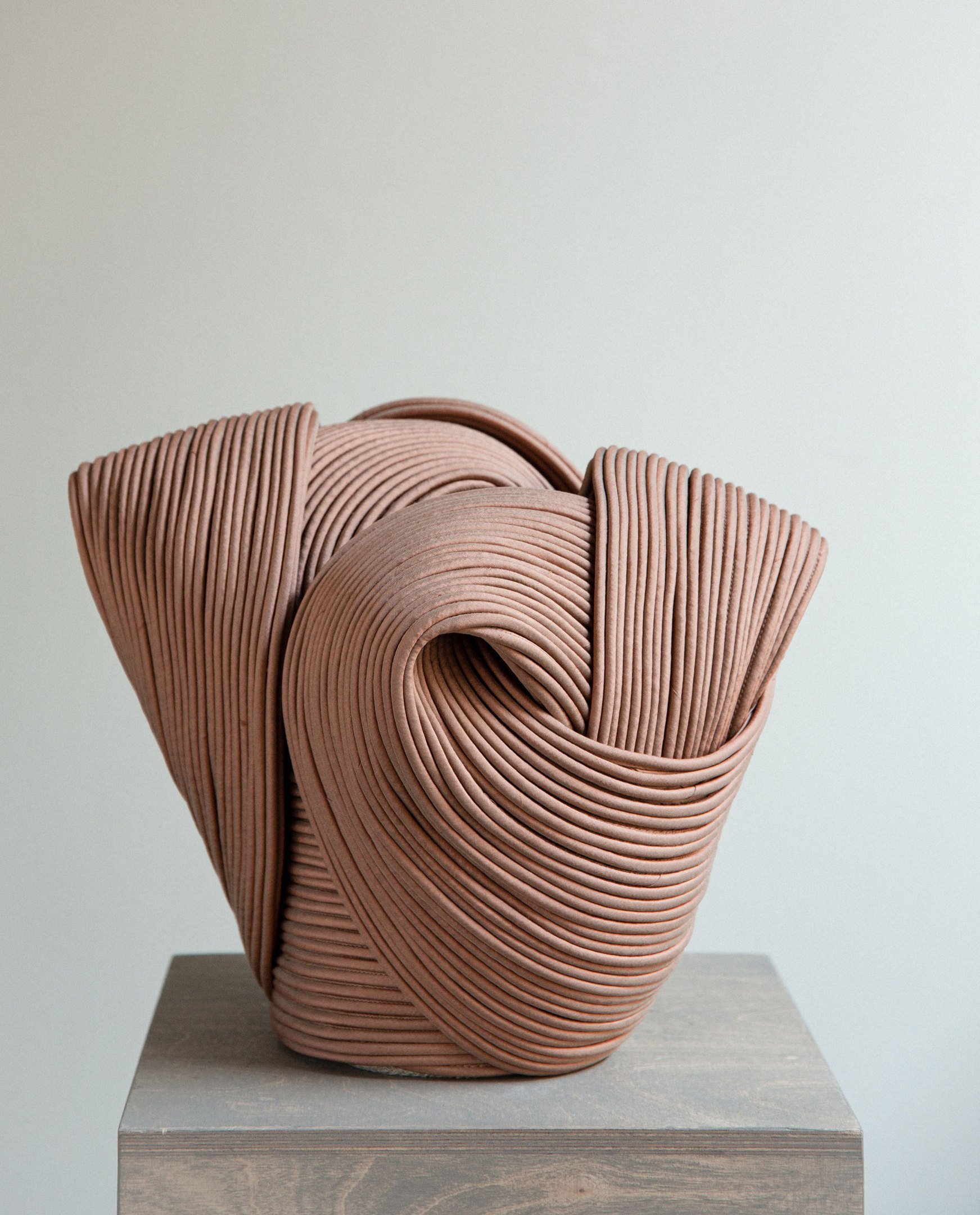 Abstract ceramic sculpture with intertwined, ribbed, terracotta-colored shapes on a wooden pedestal.