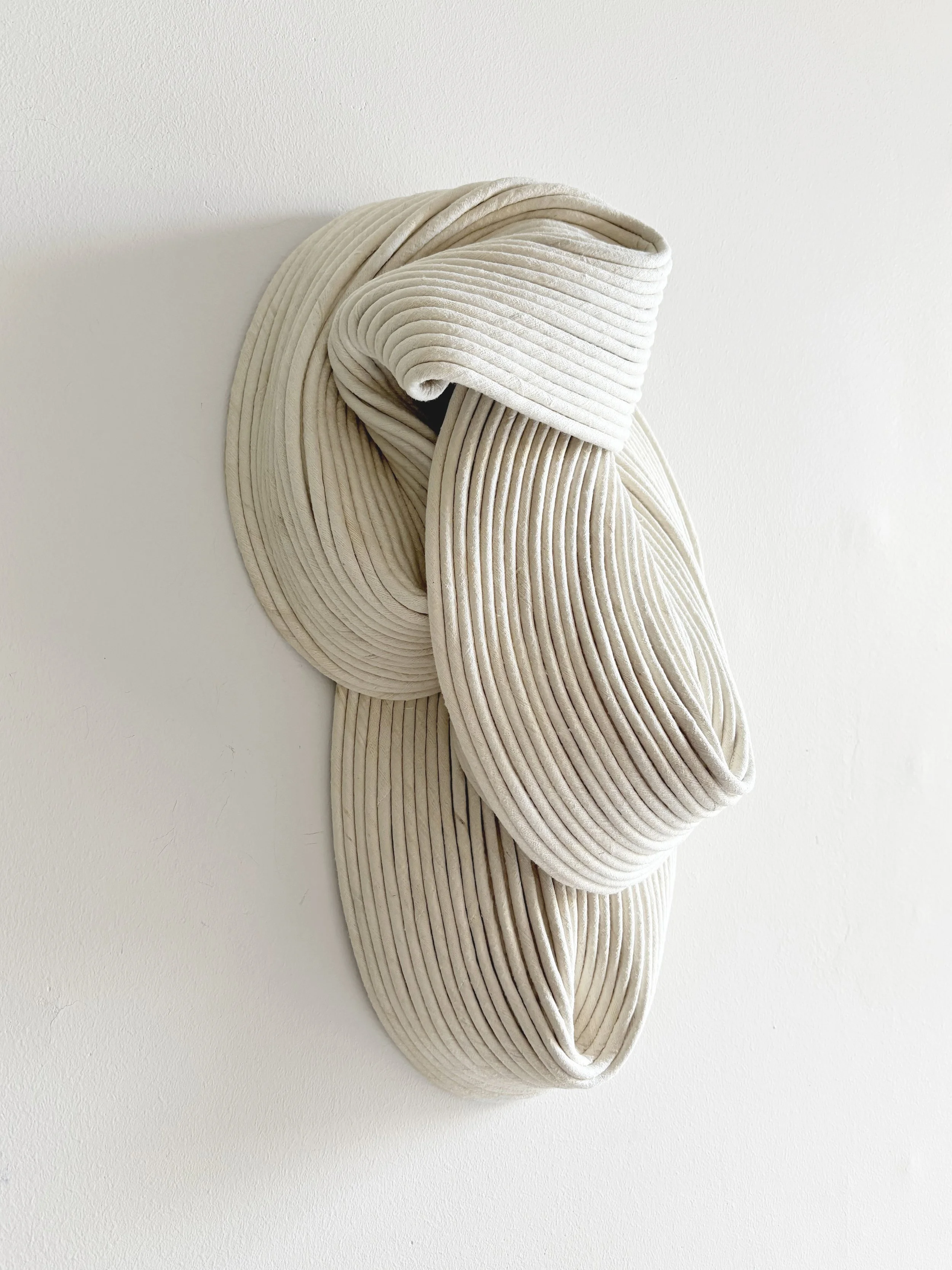A modern, abstract wall sculpture made of cream-colored, ribbed, fabric-covered loops arranged in a flowing, artistic form on a white wall.