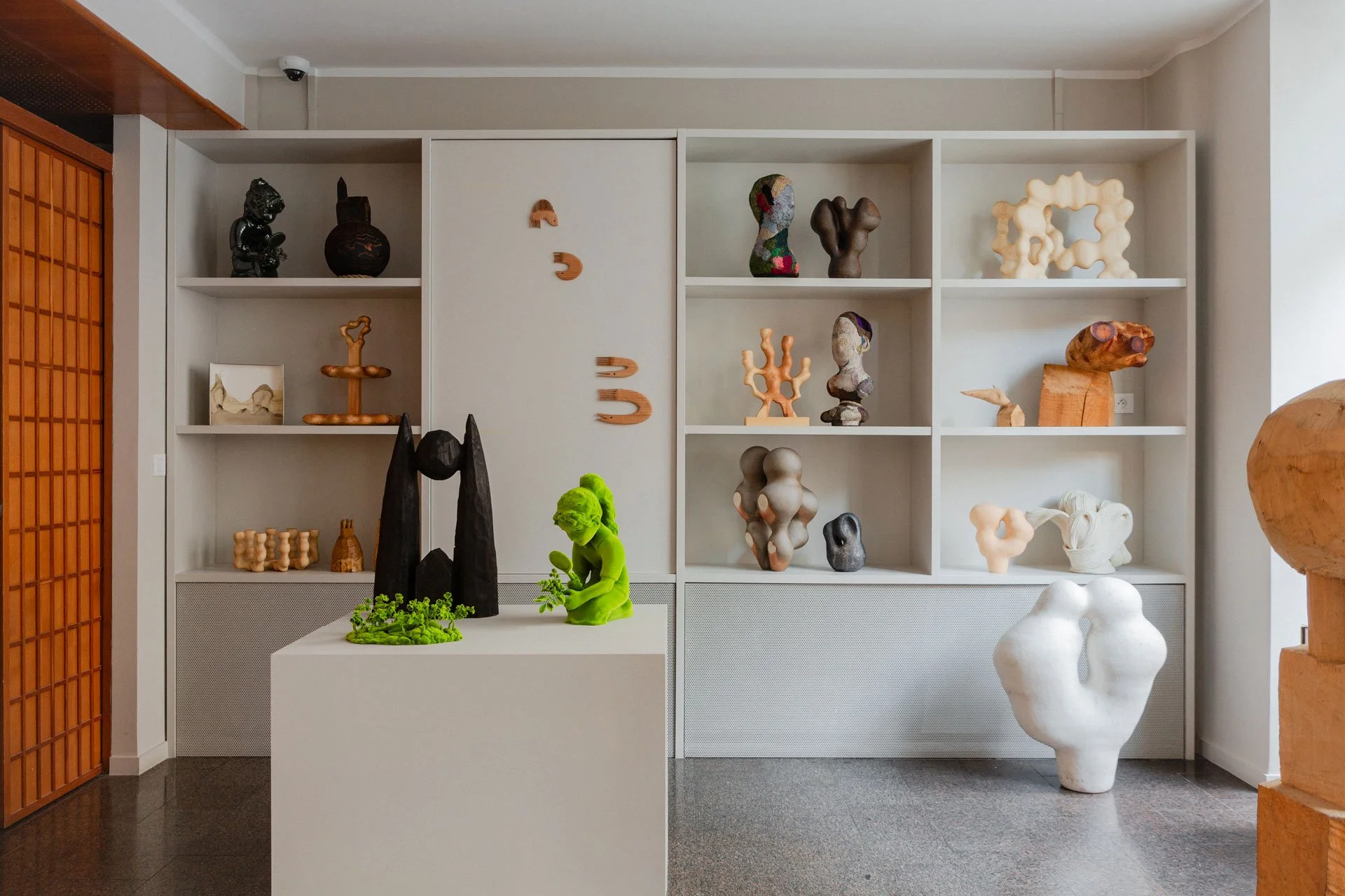 Gallery display of various modern sculptures on white shelves and on a white pedestal, with wood elements and neutral-colored walls.