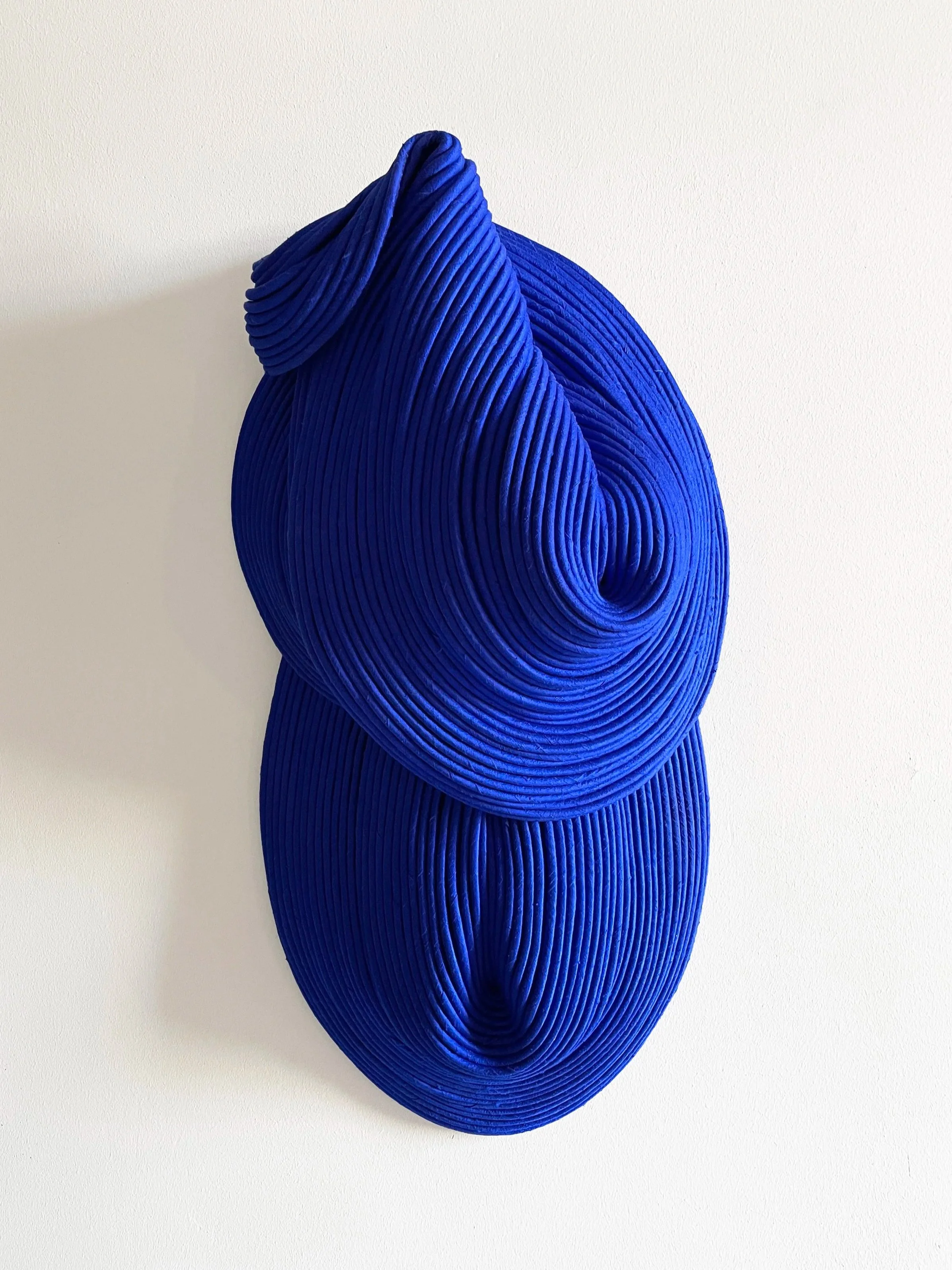 Abstract blue sculpture made of concentric, layered ridges mounted on a white wall.