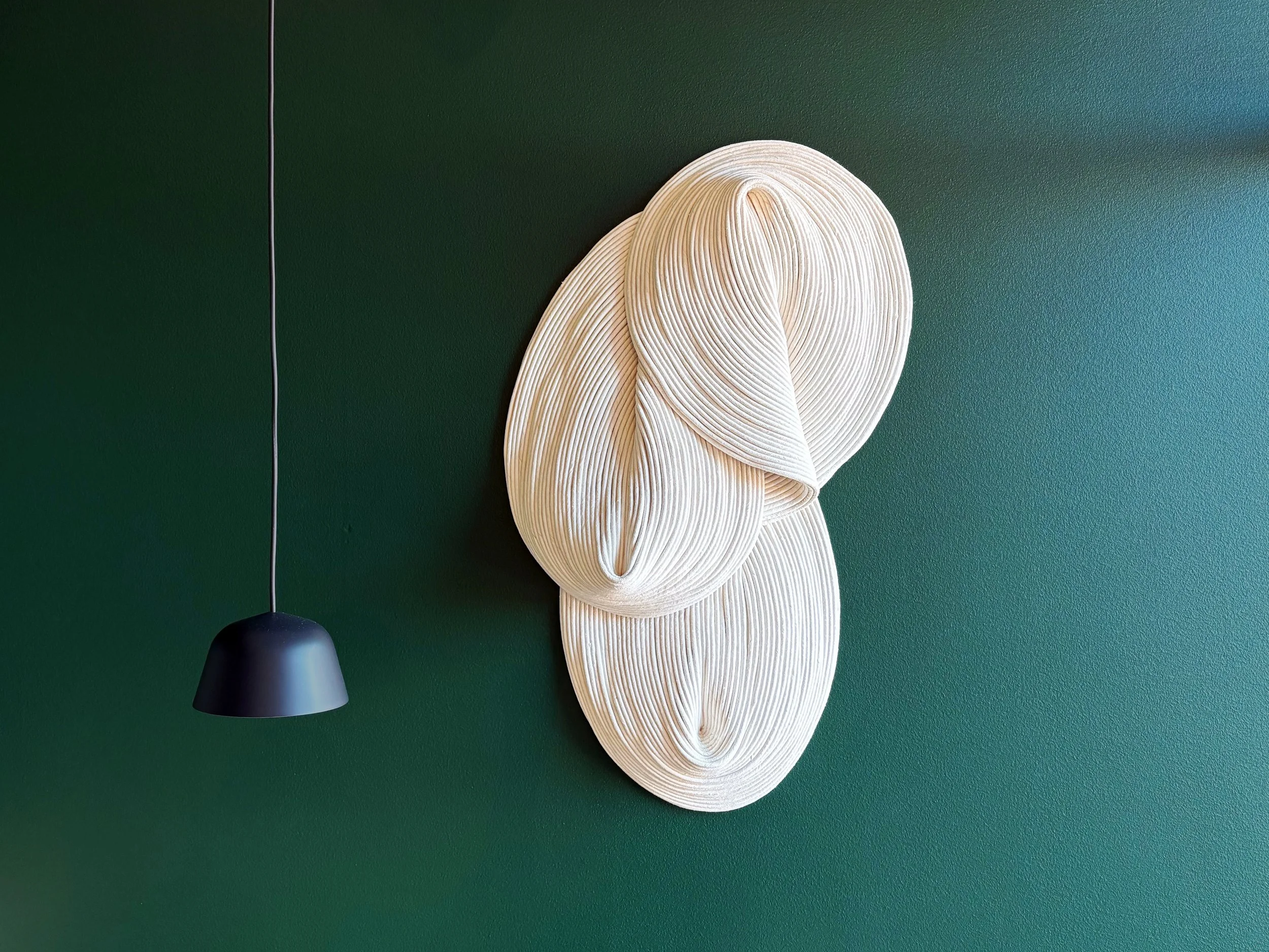 Decorative wall art with three cream-colored, swirling, layered organic shapes on a dark green wall, and a black pendant light hanging nearby.