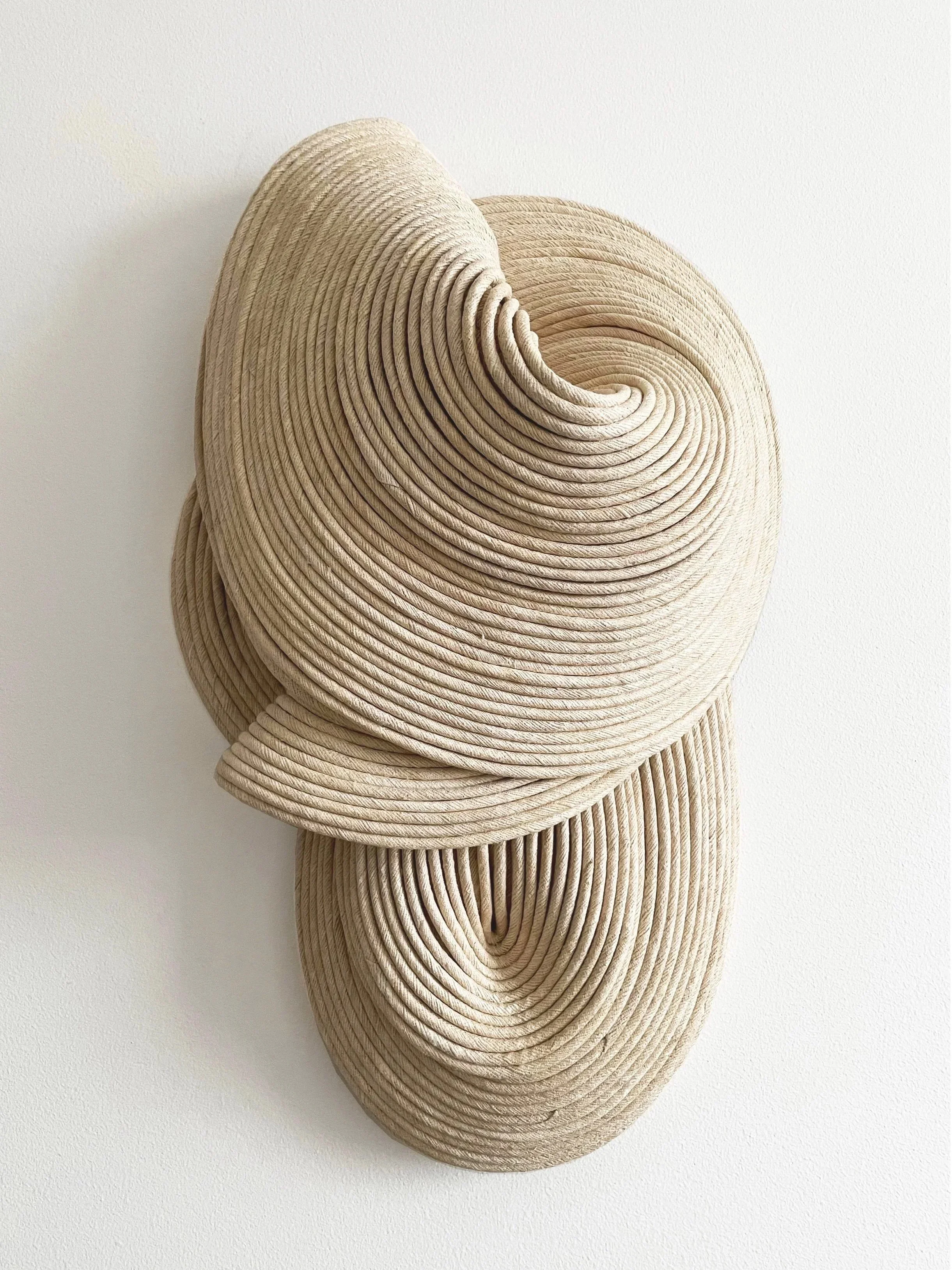 Abstract wall art made of layered, tightly coiled beige and light tan rope or cord, shaped into organic flowing forms on a plain white background.