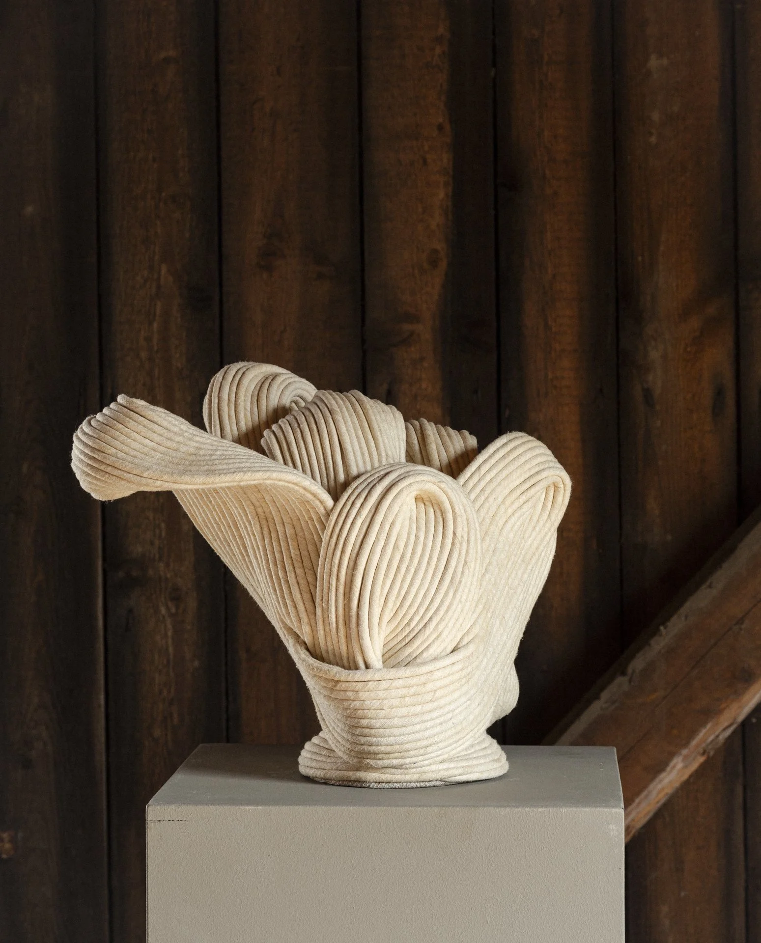 Wooden sculpture resembling a hand holding a bouquet of layered, curving wooden strips, placed on a pedestal in front of a wooden wall.