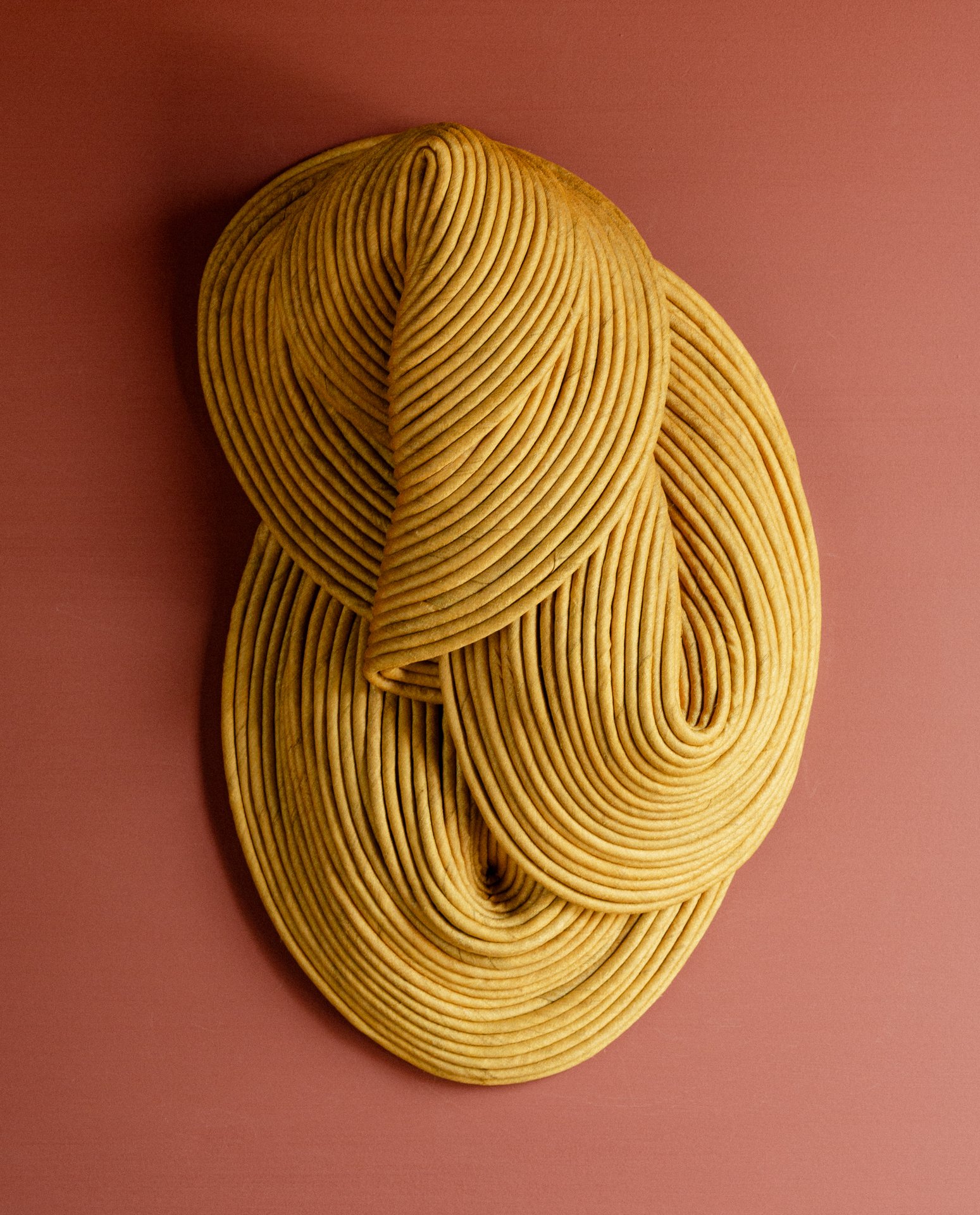 An abstract sculpture made of tightly coiled, yellowish rope arranged in an intricate, flowing pattern, mounted on a pink wall.