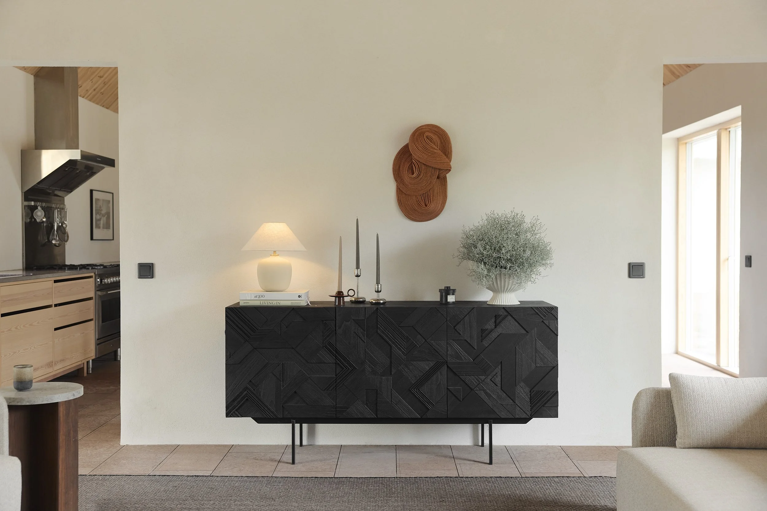 Modern living room with black sideboard, a white lamp, three long candles, a decorative vase with greenery, and wall art.