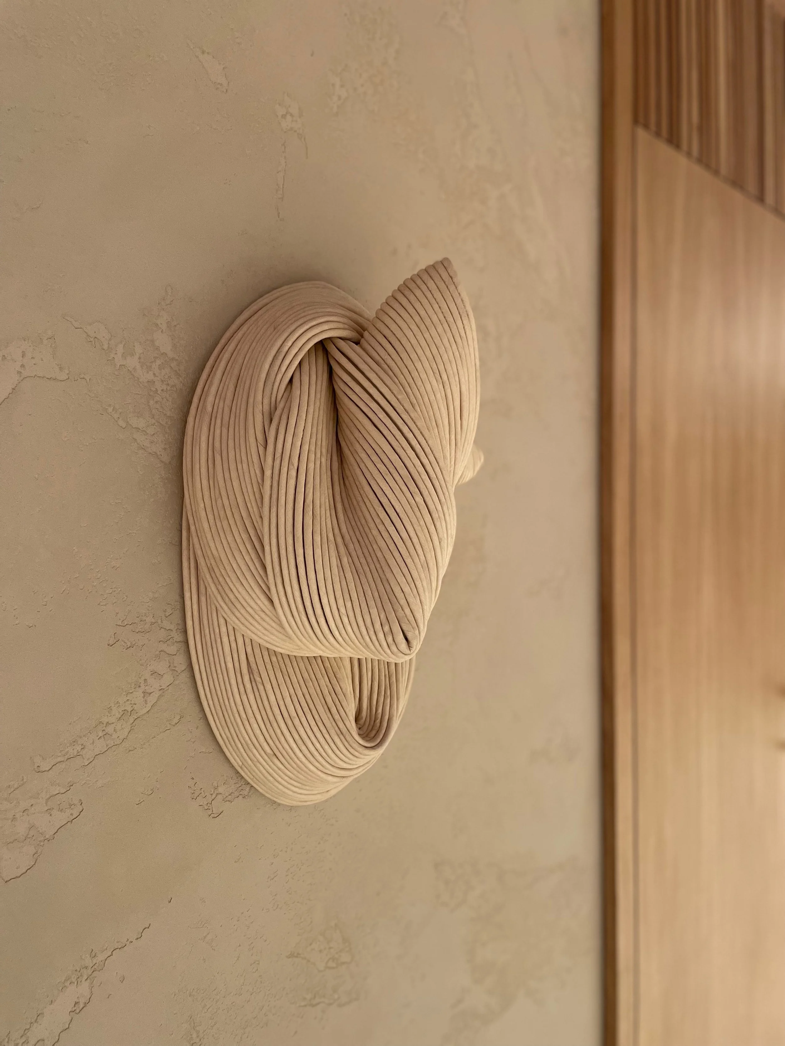 Close-up of a beige, pleated ceramic sculpture resembling a twisted, abstract form on a textured light-colored wall.