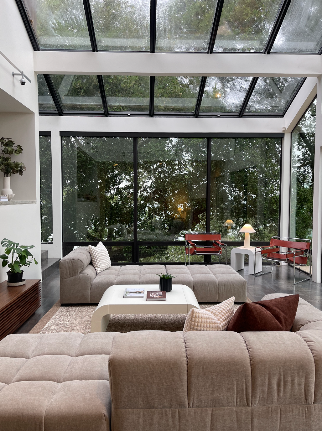 Living room with large glass windows and a skylight ceiling, beige sectional sofa, white coffee table, red chairs, small side table with a lamp, indoor plants, and a view of greenery outside.