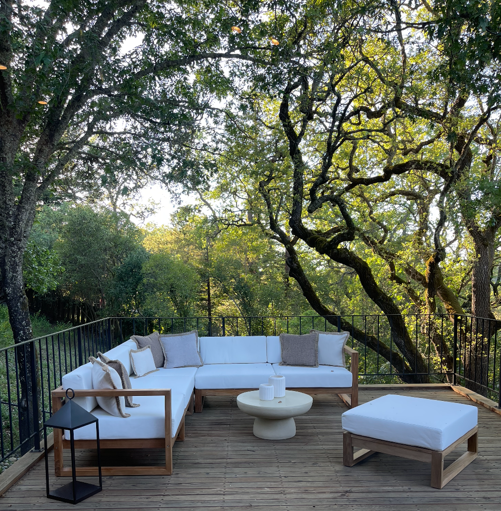 Outdoor deck with white cushioned seating, a small round coffee table, surrounded by tall trees and foliage, with natural sunlight filtering through the branches.