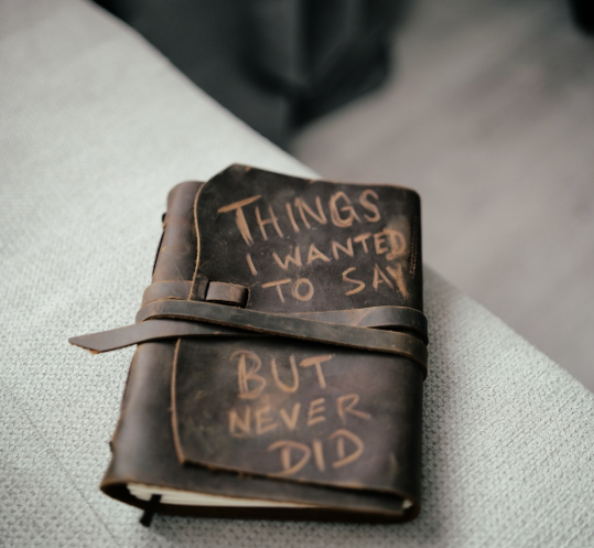 A leather journal with a strap, lying on a cushioned surface, with handwritten text reading 'Things I wanted to say but never did'.