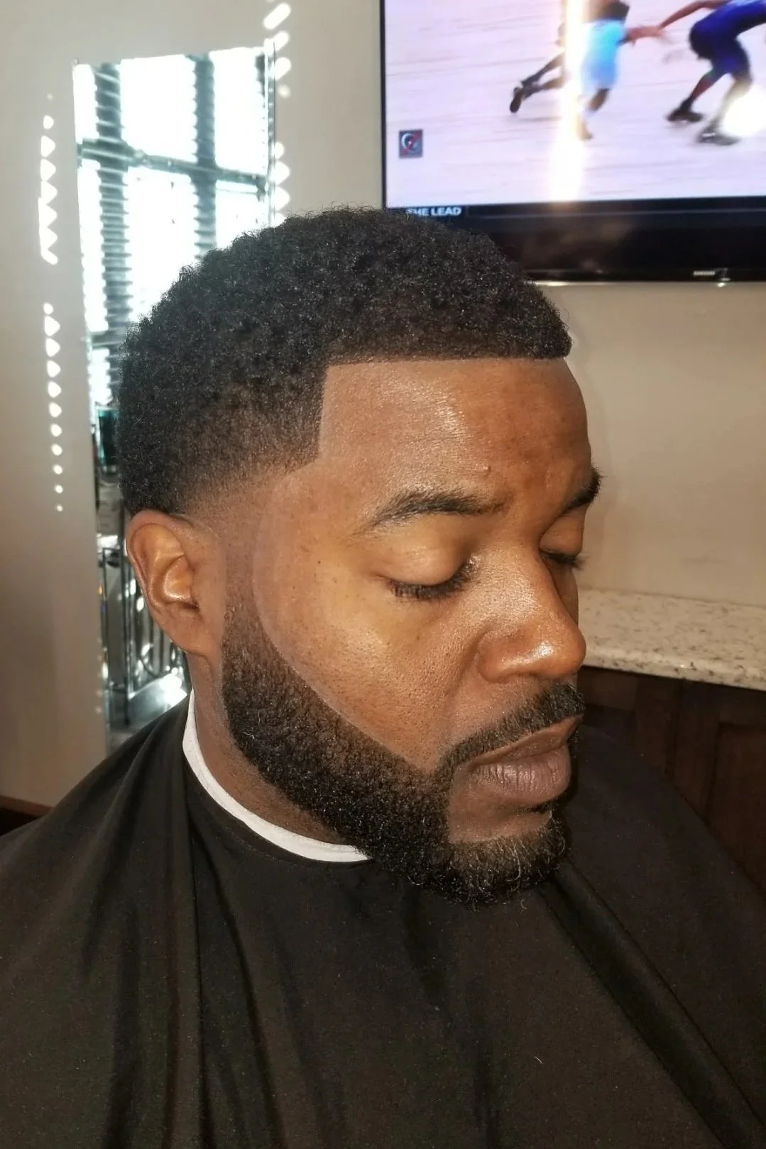 Close-up of a man with a fresh haircut and beard, eyes closed, sitting in a barber shop.
