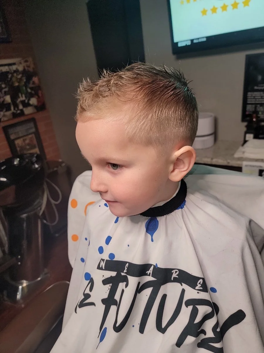 Young boy with a fresh haircut, sitting in a barber's chair wearing a cape with blue and orange paint splatter design at a barbershop