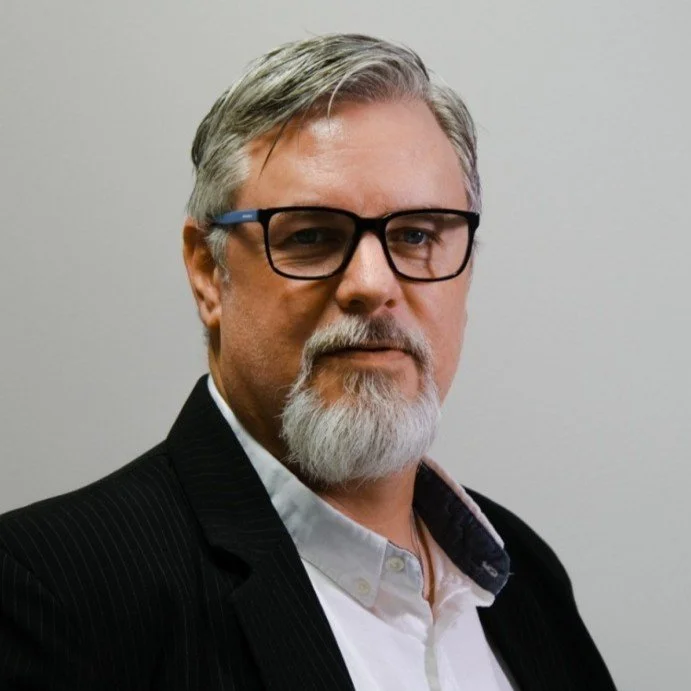 A middle-aged man with gray hair and beard, wearing glasses, a white shirt, and a black suit, standing against a plain background.