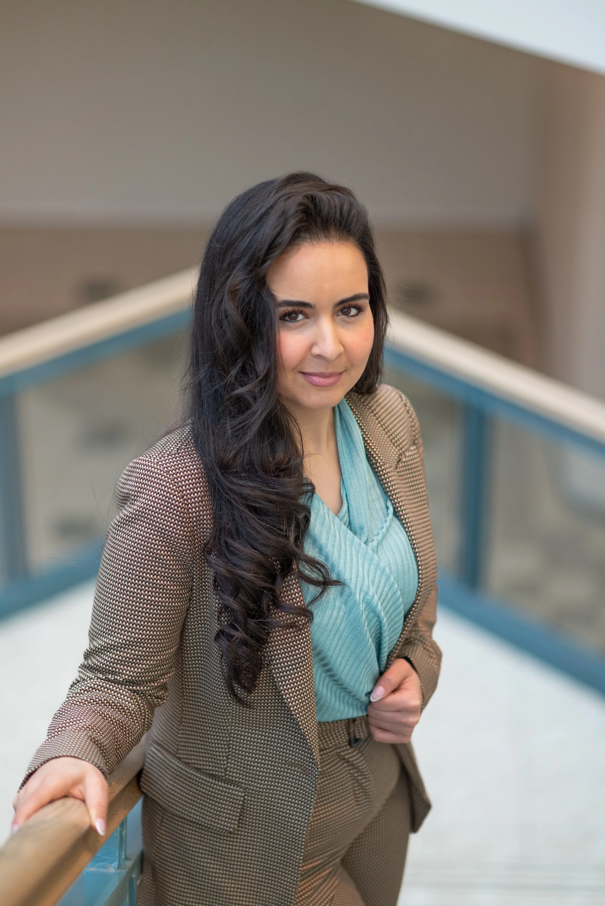 A confident woman with long wavy dark hair and light skin, dressed in a checkered suit with a turquoise top, standing on a staircase railing indoors.