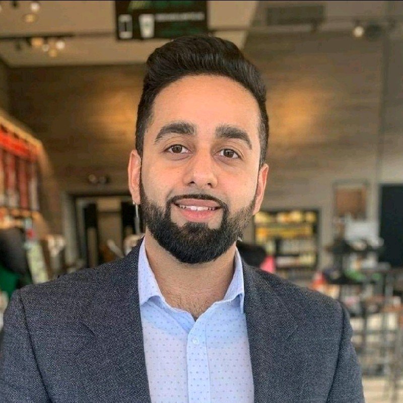 A man with dark hair and a beard smiling, wearing a light blue dress shirt and dark blazer, in a coffee shop or casual dining environment.