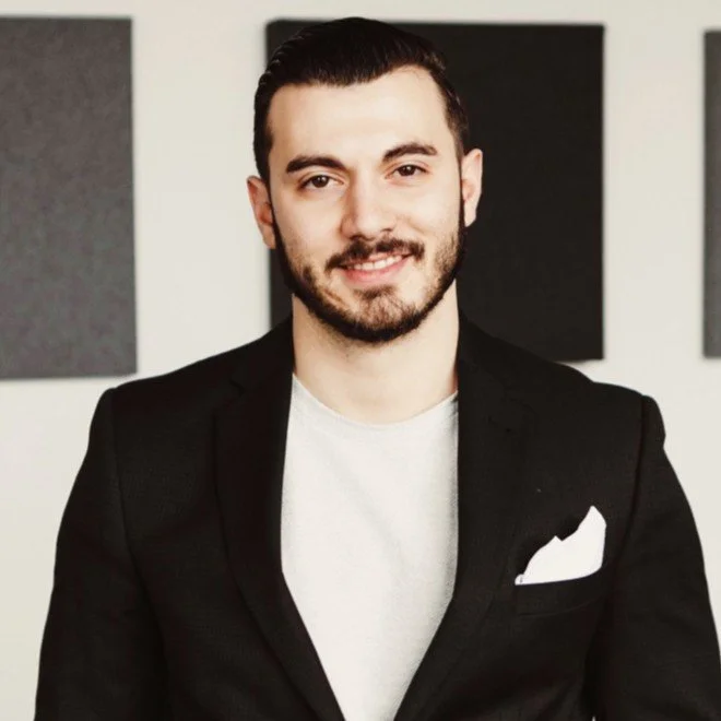 A young man with dark hair and a beard, wearing a black blazer and white t-shirt, smiling at the camera, standing in front of a wall with black and gray squares.