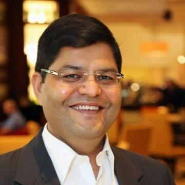 A smiling man wearing glasses and a suit in an indoor setting with warm lighting