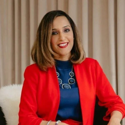 A woman with shoulder-length brown hair, wearing a red blazer over a blue top, sitting in front of beige curtains, smiling at the camera.