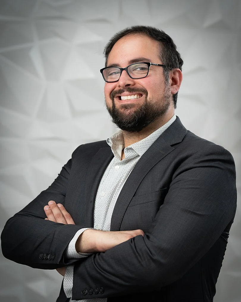 A smiling man with glasses, a beard, and dark hair wearing a suit and crossed arms, standing in front of a textured white background.