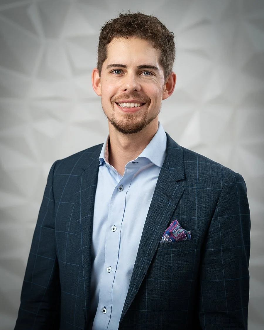 A young man with short, curly brown hair and a beard, wearing a navy blue checked blazer, light blue shirt, and a colorful pocket square, smiling at the camera against a gray geometric background.